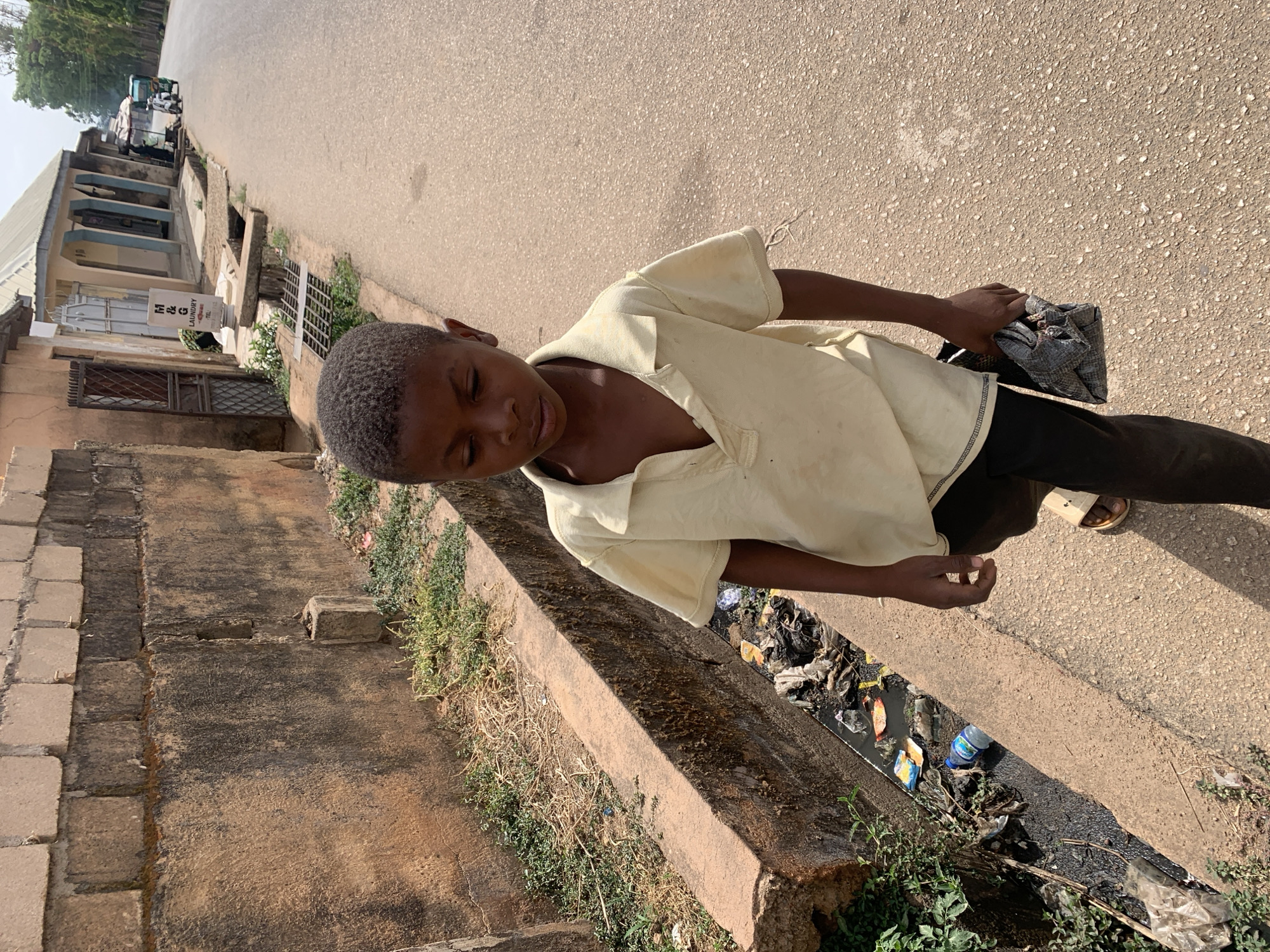 A boy stands on the edge of a street in Jos, Nigeria. He wears a light yellow, short-sleeved shirt and dark pants, and sandals. He holds a gray cloth or bag in his right hand. To his left is a concrete curb bordering a drainage ditch filled with trash. Beyond the ditch, a brick building rises. The street stretches into the distance, where vehicles are visible. The sky is overcast.