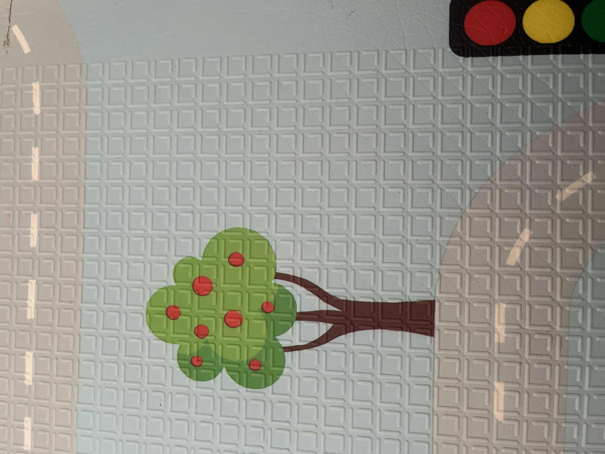 This is a close-up, top-down media file of a play mat designed for children. The mat features a street scene with a textured, light blue background overlaid with a grid pattern. In the foreground, a stylized green tree with red circular fruit is depicted. The tree has brown branches extending to the right. To the right of the tree, a portion of a traffic light is visible, showing red, yellow, and green circular lights against a black background. The road is indicated by grey areas with white dashed lines. The scene is brightly lit, suggesting daytime, with no discernible weather conditions. There is no visible text. The overall impression is a safe, indoor play environment. The location context provided is Chelyabinsk, Russia, which is not visually represented in the image itself.