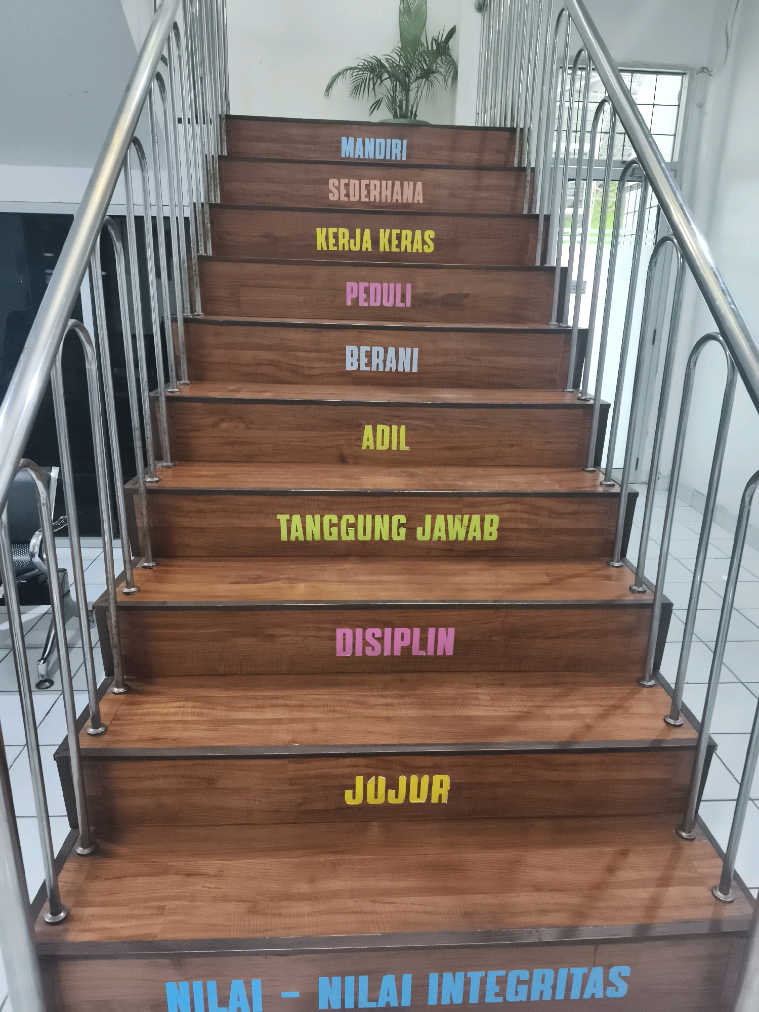 The image shows a wooden staircase with metal railings, located indoors. Each step of the staircase has a word written on it in colored block letters, presumably representing values or principles. From top to bottom, the words are: MANDIRI (blue), SEDERHANA (peach), KERJA KERAS (yellow), PEDULI (pink), BERANI (light blue), ADIL (green), TANGGUNG JAWAB (lime green), DISIPLIN (pink), JUJUR (yellow), and at the bottom, NILAI - NILAI INTEGRITAS (light blue). The environment appears to be a well-lit interior space, possibly an office or educational institution, with a plant visible at the top of the stairs. There are no people visible in the image. The staircase is clean and well-maintained. The location is identified as Unknown, Indonesia.