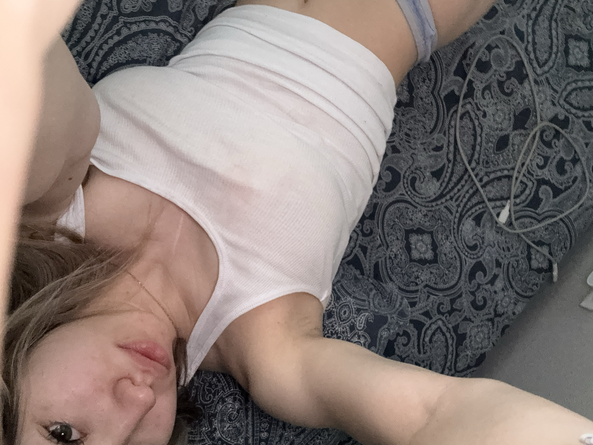A woman lies on a paisley-patterned bed, wearing a white tank top. Her face is close to the camera. A pair of headphones are on the bed next to her. The location is not discernible from the image.