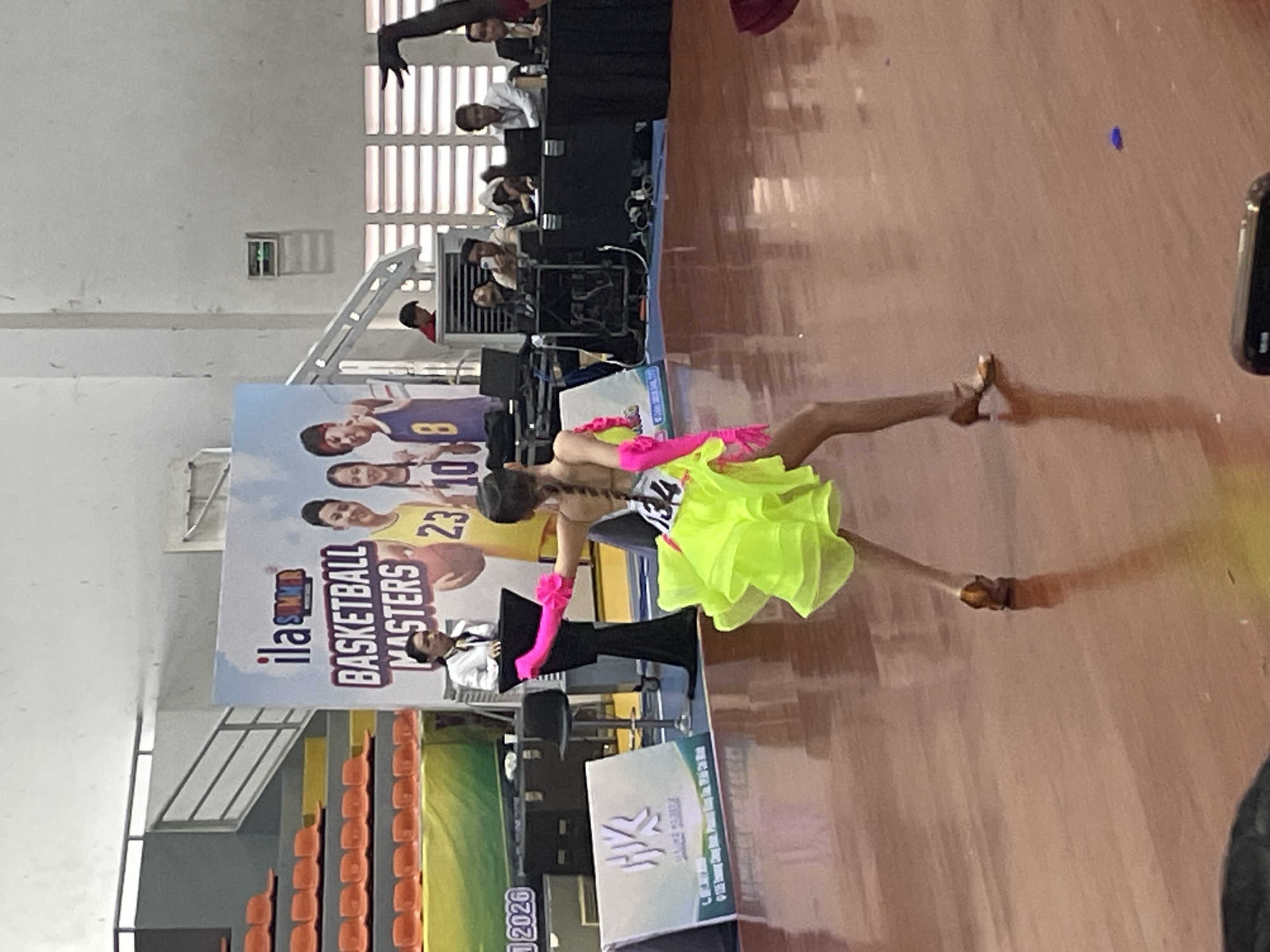 In the city of Thuận An, Vietnam, within what appears to be an indoor sports arena, a young female dancer is captured mid-performance. She is wearing a vibrant neon yellow costume with ruffled layers and bright pink arm coverings, and has the number "134" visible on her back. Her stance and the dynamic pose suggest she is engaged in a competitive dance event.
The background features a large banner with the text "ila SUMMER BASKETBALL MASTERS" and images of basketball players. Several other individuals are present in the background, including a woman in a white collared shirt standing behind a podium, likely an official or announcer. Other people are seated at tables with laptops and appear to be observing or working at the event. The flooring is a polished wooden surface, reflecting the lighting and the dancer. Orange stadium seating is visible in the upper left of the frame, indicating a spectator area. The overall environment suggests a formal event, possibly a competition or exhibition.