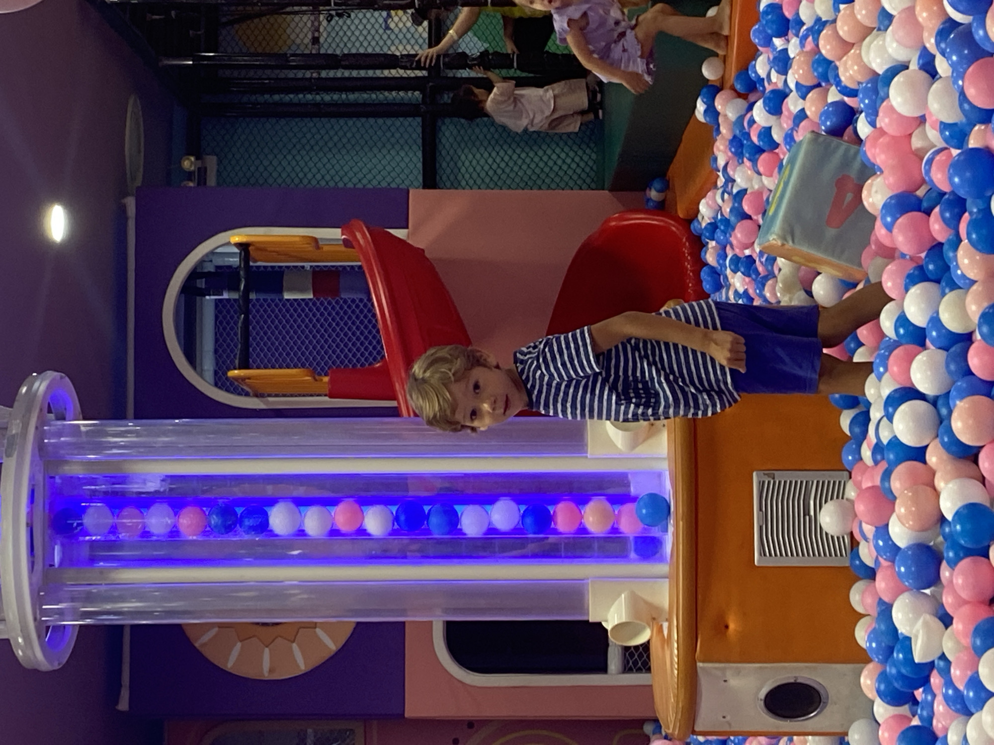 In the city of Phu Thuy, Vietnam, a young boy with blond hair stands in the foreground of a brightly colored indoor playground. He is wearing a blue and white striped t-shirt and blue shorts, and is looking directly at the camera with a slight smile. Behind him, a tall, clear tube filled with pink, blue, and white balls is illuminated by purple lights. The playground features a red slide in the middle ground and a ball pit filled with numerous pink, blue, and white balls in the foreground and extending to the right. Other children are visible in the background, playing in the playground. The environment is designed for children's recreation, with soft surfaces and playful structures.