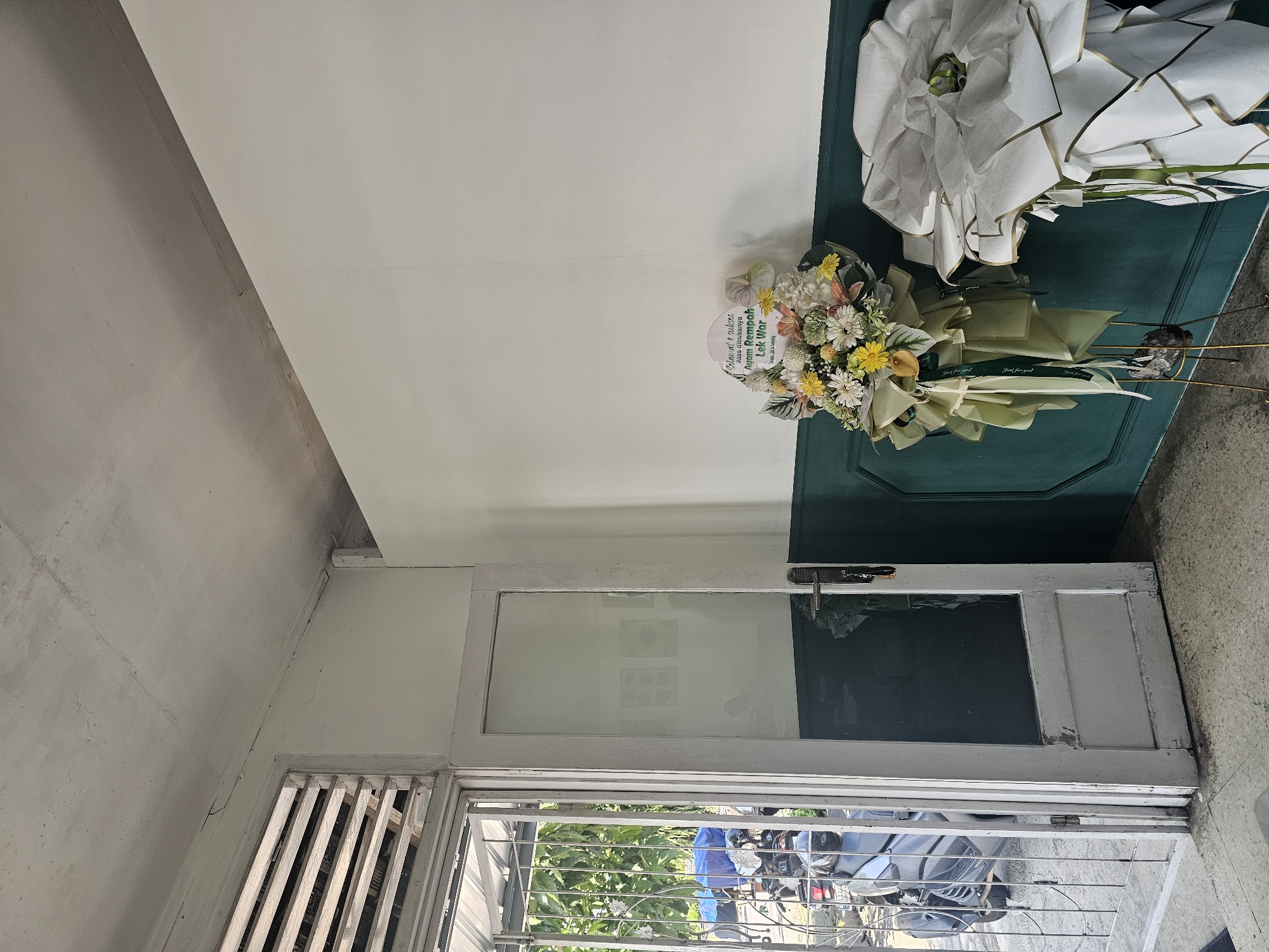 The image shows the interior of a building, likely a shop or restaurant, in Banyuurip, Indonesia. The room is sparsely furnished, with white walls and a dark green wainscoting. A white-painted door with a glass pane is open to the left, revealing a glimpse of the outdoors with foliage and a parked motorcycle. In the foreground, two flower arrangements stand on display. The one on the left is a bouquet of various flowers, including yellow, white, and peach-colored blooms, adorned with green ribbon and a white circular sign with green text that reads "Selamat & Sukses Atas Dibukanya Ayam Rempah Lek War". The arrangement on the right is a larger, more elaborate display wrapped in white paper with gold trim. The overall impression is one of a newly opened establishment, possibly a food business, as suggested by the flower arrangement's inscription. The lighting is natural, casting soft shadows. The floor is tiled in a light color with some dark staining. The ceiling shows some visible cracks and imperfections.