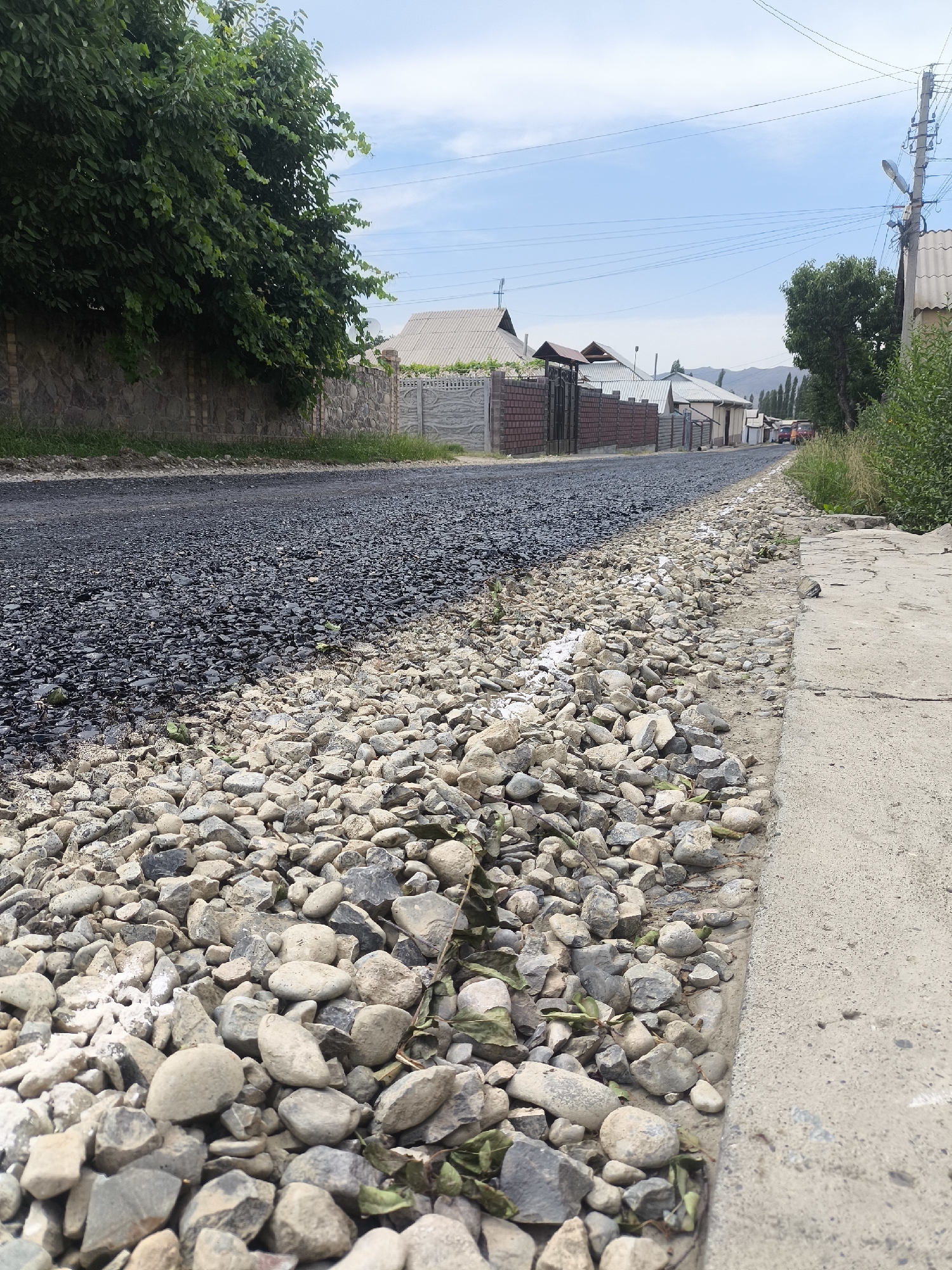 The image depicts a street in the city of Kyzyl-Kyya, Kyrgyzstan, with a section of the road under construction or repair. The foreground is dominated by a curb made of rough concrete, bordering a wide expanse of loose, light-colored gravel. Adjacent to the gravel is a newly laid layer of dark asphalt, stretching into the middle ground. The asphalt appears to be a rough, uncompacted surface, suggesting recent application.
Beyond the road, a stone wall with a brick coping lines the left side of the image, topped by dense green foliage. Across the street, several houses are visible, characterized by their varied rooflines and fencing. The architecture suggests a residential area. A utility pole with numerous wires stands on the right side of the road, indicating the presence of public services. The sky is a pale blue with scattered clouds. In the distance, more houses and trees are visible, leading towards a mountainous or hilly landscape. The overall impression is of a semi-urban or suburban environment undergoing roadwork. No people are visible in the scene.