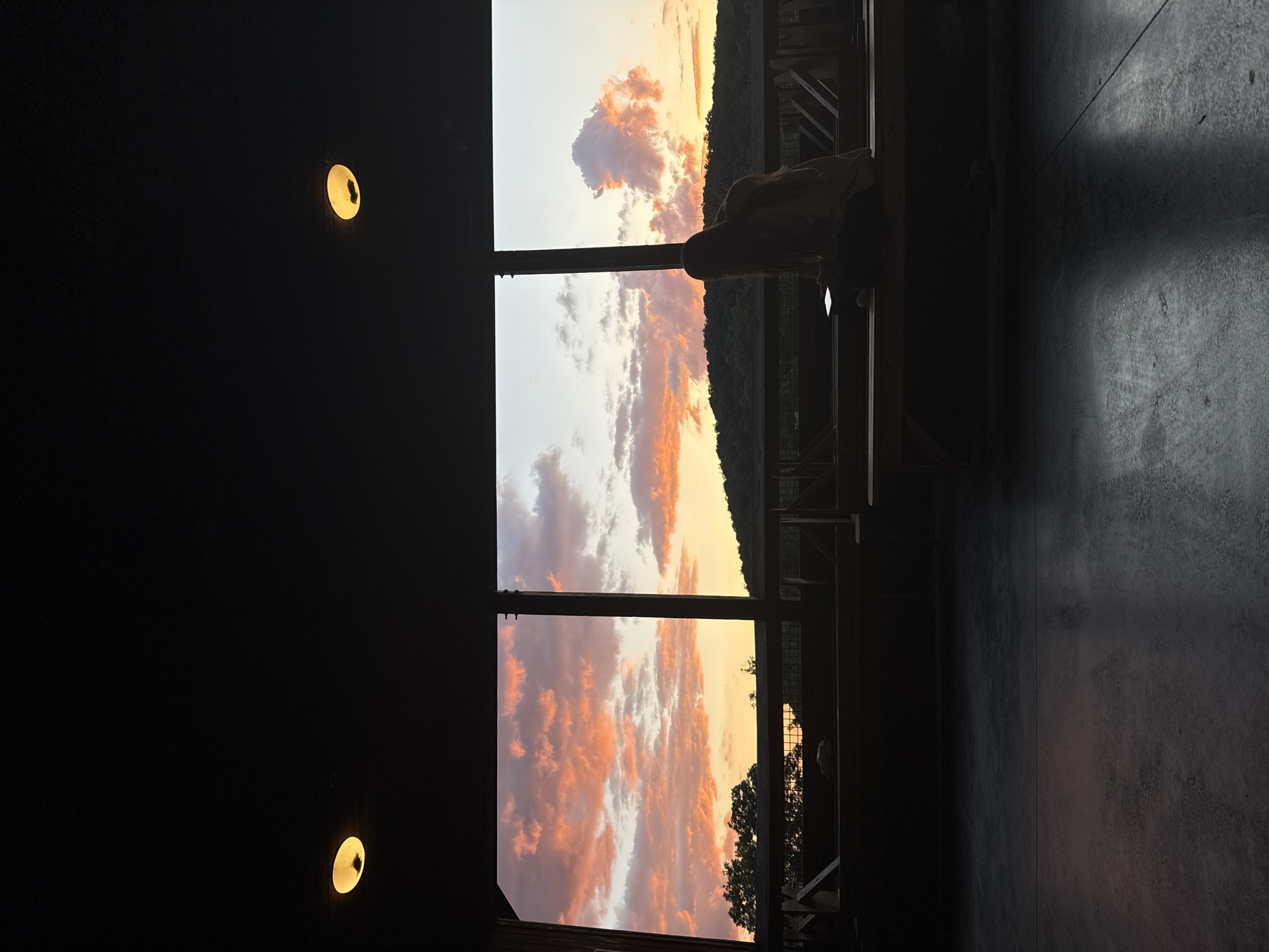An outdoor scene is captured at dusk, likely in Washington Township, United States of America. The foreground is a dark, concrete floor, reflecting some of the ambient light. Above this is a dark ceiling with two circular light fixtures, one on the left and one on the right, emitting a warm yellow glow.
The middle ground consists of a covered outdoor seating area with wooden benches and railings. The area is largely in shadow, but the edges of wooden structures are visible.
The background opens up to a panoramic view of a sunset through a large, open frame. The sky is filled with dramatic clouds painted in vibrant hues of orange, pink, and yellow, indicating the sun is low on the horizon. Silhouetted against this colorful sky are trees on the left and a gentle, dark hill or mountain range in the distance.
A single person, silhouetted from behind, is seated on a wooden bench in the middle ground, facing the sunset. They appear to be looking down, possibly at a phone. The person is wearing a hooded jacket. Their posture suggests a moment of quiet contemplation or engagement with a personal device while observing the natural spectacle. The overall atmosphere is serene and peaceful.