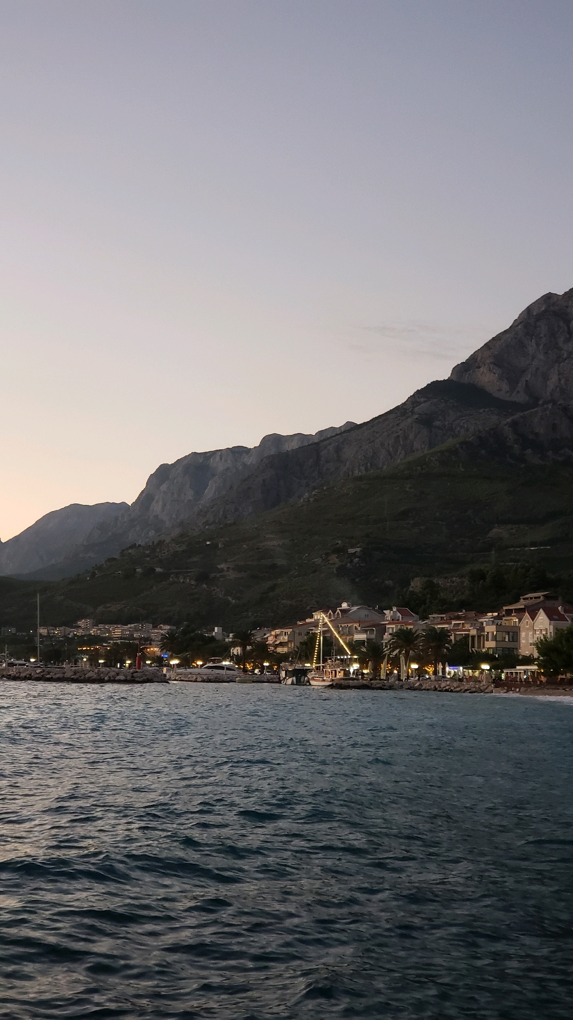 The image shows a coastal scene at dusk. A body of dark, rippling water occupies the foreground. In the mid-ground, a town is nestled along the coastline. Buildings with illuminated windows and lights are visible, suggesting it is a developed area with infrastructure. Several boats, including yachts and a larger vessel with decorative lights, are docked or anchored in the harbor. Behind the town, steep, dark, and rugged mountains rise, their slopes covered in dense vegetation. The sky above is a pale gradient, transitioning from a lighter tone near the horizon to a darker shade at the top, indicative of twilight. The scene is set in Općina Tučepi, Croatia, a location known for its coastal landscape and mountainous backdrop. There are no discernible people in the image.