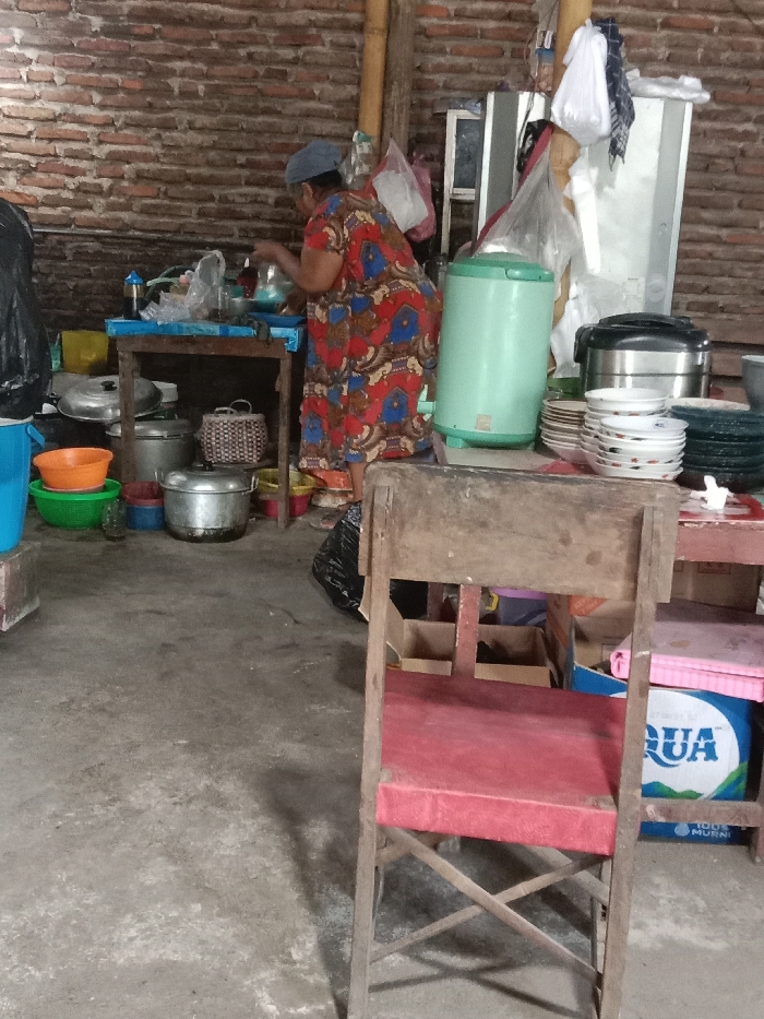 A woman in a red and blue dress cooks at a table in a rustic kitchen. Behind her are various pots, pans, and appliances. A light green water dispenser sits next to stacks of plates. In the foreground, a worn wooden chair with a red cushion is visible. An Aqua brand water bottle is partially visible behind the chair. The kitchen's background consists of an unfinished brick wall.