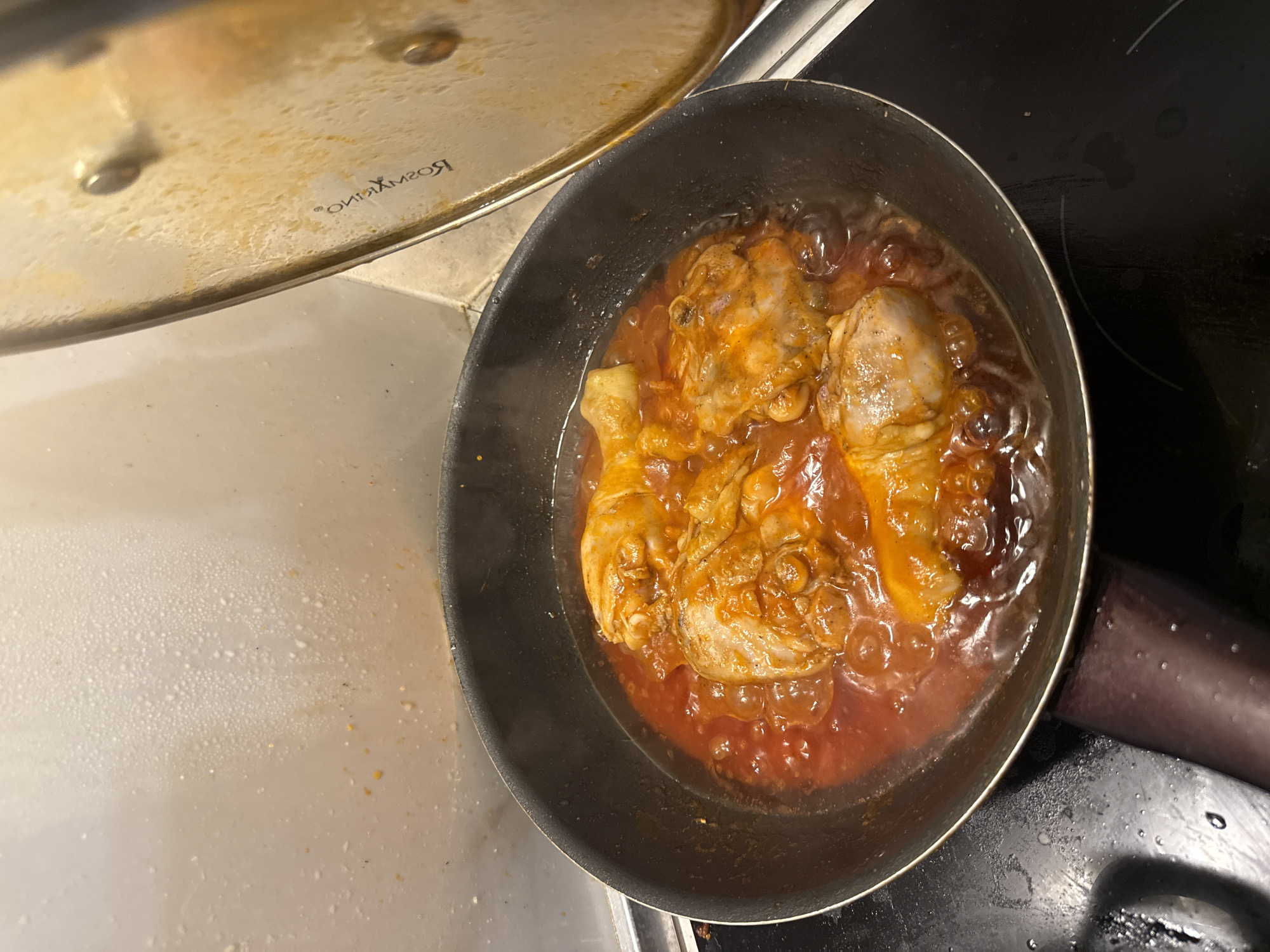 The image shows a close-up, overhead view of chicken legs cooking in a red sauce within a black frying pan. The pan is situated on a black stovetop, with a partially opened silver lid to the right of the pan. The lid has some cooking splatters on it and a logo that appears to read "ROSMARINO". The background consists of a white tiled backsplash with visible moisture and some food debris. The stovetop also shows water droplets and some dark smudges. The scene appears to be an indoor kitchen environment. There are no people or specific landmarks to indicate a location within Boreti, Montenegro.