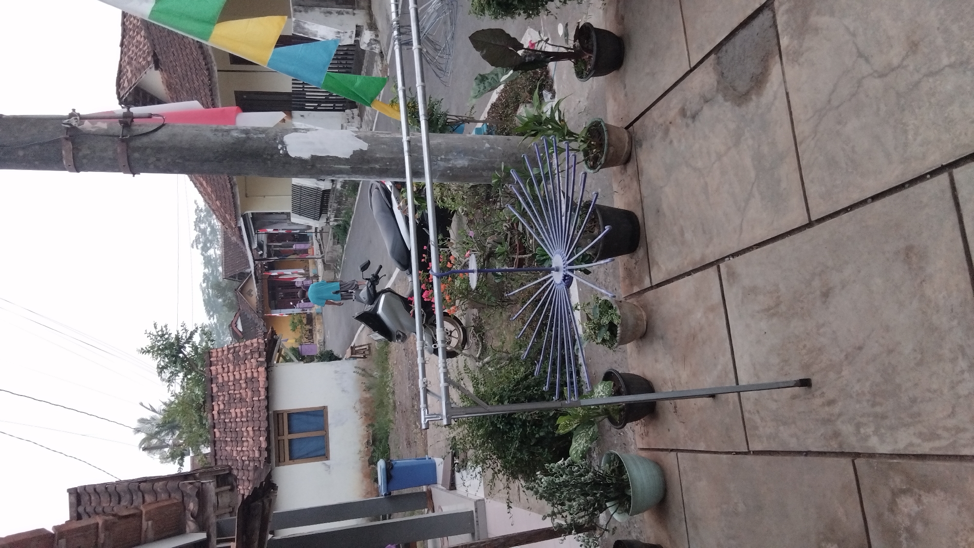A street in Tulis, Indonesia, features houses with terracotta roofs. In the foreground, potted plants sit on a paved patio. A clothes drying rack is positioned in front of the plants. Behind the rack, a motorcycle is parked. A man in a blue shirt walks away from the viewer on the street. A utility pole with Indonesian flags is visible on the right side of the image. The sky is overcast.