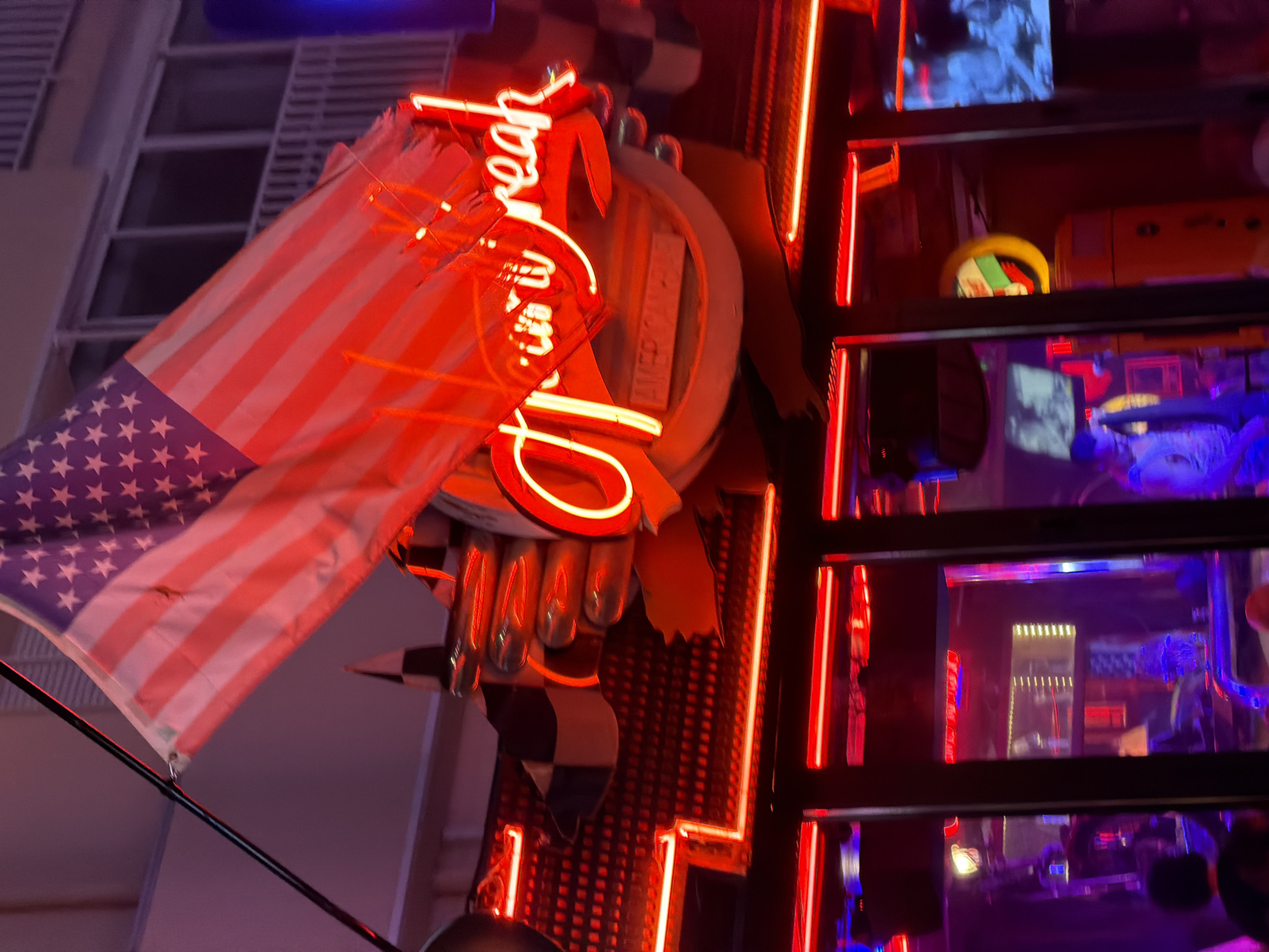 The image depicts the exterior of a bar or restaurant in Benidorm, Spain, characterized by bright red neon lighting. An American flag is prominently displayed, partially obscuring a neon sign that appears to read "TIN U BREAK". The architecture suggests a building façade with multiple floors and windows. Inside, through the darkened windows, glimpses of television screens and what seem to be patrons can be observed, all bathed in the pervasive red glow. The overall atmosphere is that of a lively nighttime establishment.