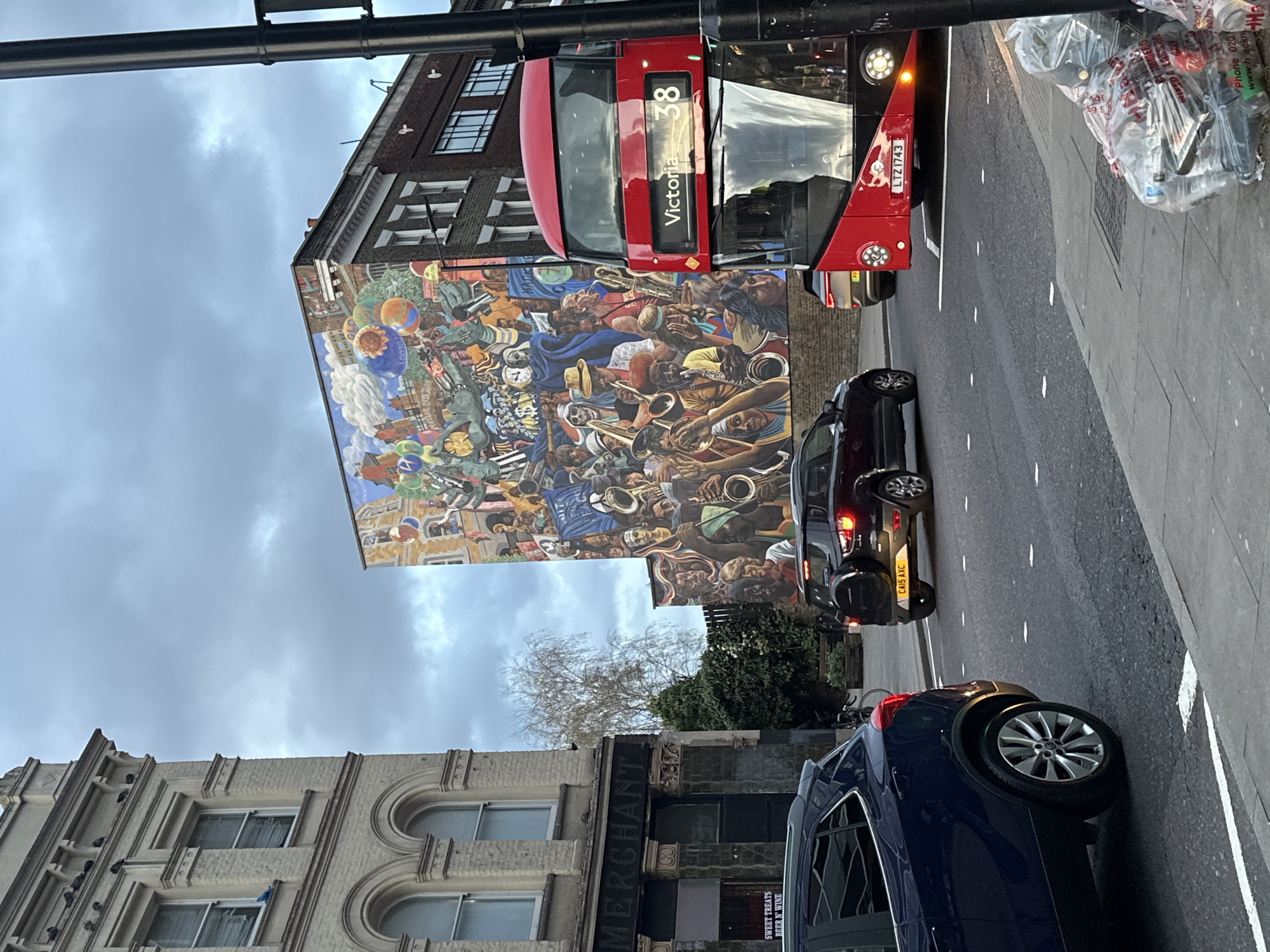The image captures a street scene in London, United Kingdom. A red double-decker bus with the route "38 Victoria" prominently displayed is partially visible at the top. Adjacent to the bus, a large, detailed mural adorns the side of a building, featuring numerous figures, instruments, and various design elements.
Below the bus and mural, vehicles are parked along the roadside, including a dark-colored car and a blue car. A street sign post is visible at the top left corner of the image.
Buildings line the street, one of which has a sign that reads "MERCHANT" with the additional text "SWEET TREATS BEER N' WINE" visible below. The sky above is overcast with clouds. There is a plastic bag filled with trash. The license plate CA15 AXC is visible. Also visible is license plate LTZ 1743.