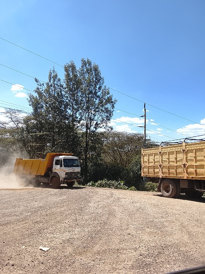 The image depicts an outdoor environment in Kiambu Township ward, Kenya, under a clear blue sky with scattered white cumulus clouds. The ground is a light brown gravel or dirt surface, with a small crumpled white object visible in the lower-left foreground. A white-cab, yellow-bed dump truck is positioned in the midground on the left, oriented towards the right, generating a prominent dust cloud around its rear wheels, indicating movement. To the right, the rear section of a second, stationary truck with a light-brown wooden cargo bed is visible. The background consists of dense green foliage, including tall trees and shrubs. Multiple horizontal power lines traverse the upper portion of the frame, and a utility pole with insulators is present in the upper-right quadrant. No people are visible.