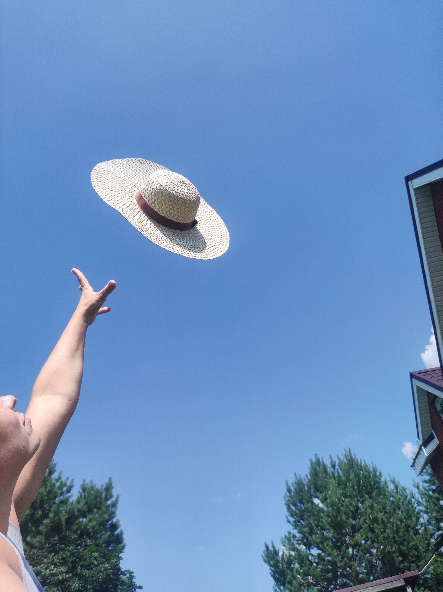A person's arm is raised, with their hand reaching towards a straw hat that is suspended in the air against a clear blue sky. The hat is light-colored with a darker band around its crown. In the background, there are green trees and the corner of a red-roofed building. The image appears to be taken from a low angle, looking up. The location is the city of Фоки, Russia.