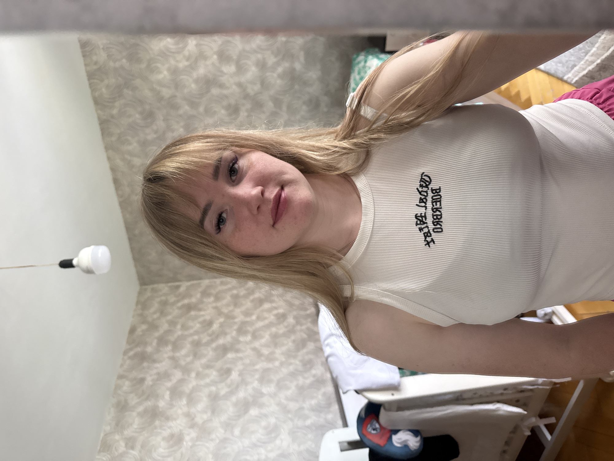 A fair-skinned woman with blond hair and bangs is centered in the image, wearing a white ribbed tank top with black lettering, likely in a residential indoor environment. The background shows patterned wallpaper on the left, a partially visible bed with a pink blanket, and various objects suggesting a bedroom or living space. An obscured light fixture is visible at the upper left. The location is Марковский, Russia.