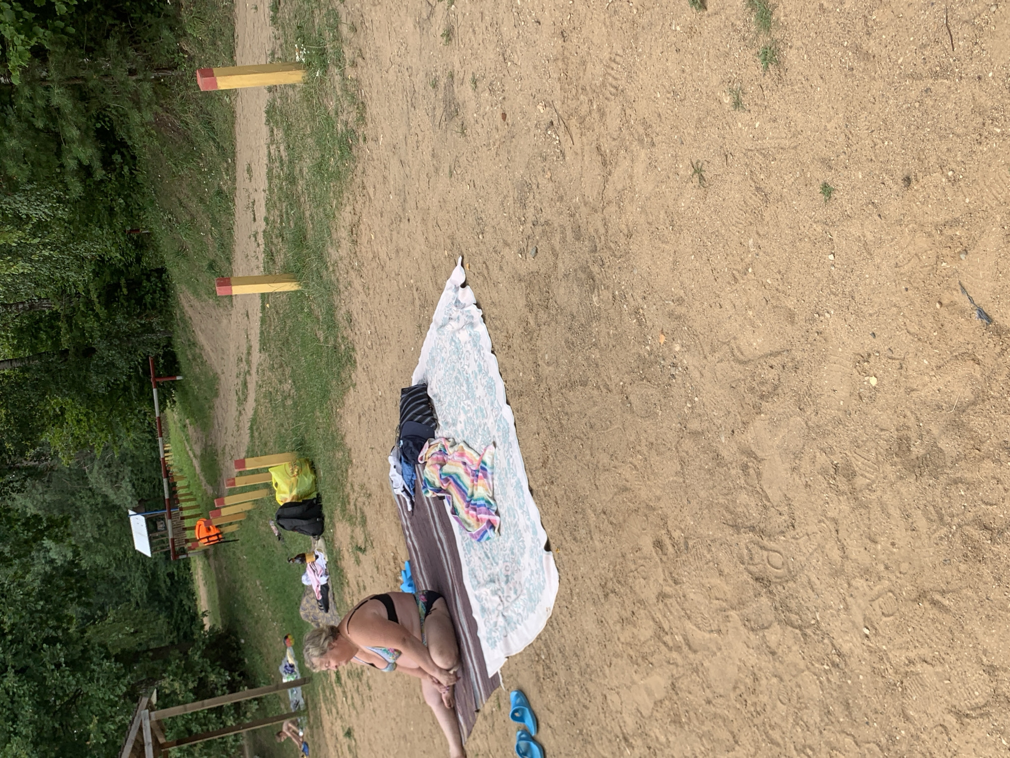 In the city of Грозаўскі сельскі Савет, Belarus, a woman in a bikini sits on a blanket on a sandy area, facing left. She has light skin and blonde hair. Beside her on the blanket are several items of clothing, including a striped towel and a colorful patterned garment. A black backpack and a yellow bag are also present. In the background, a wooden structure with a roof is visible, along with trees and a grassy slope. A line of yellow posts with red tops runs along the edge of a path. Further back, an orange life jacket is hanging on one of the posts. Blue flip-flops are on the sand to the left of the blanket. The ground is sandy with patches of grass.