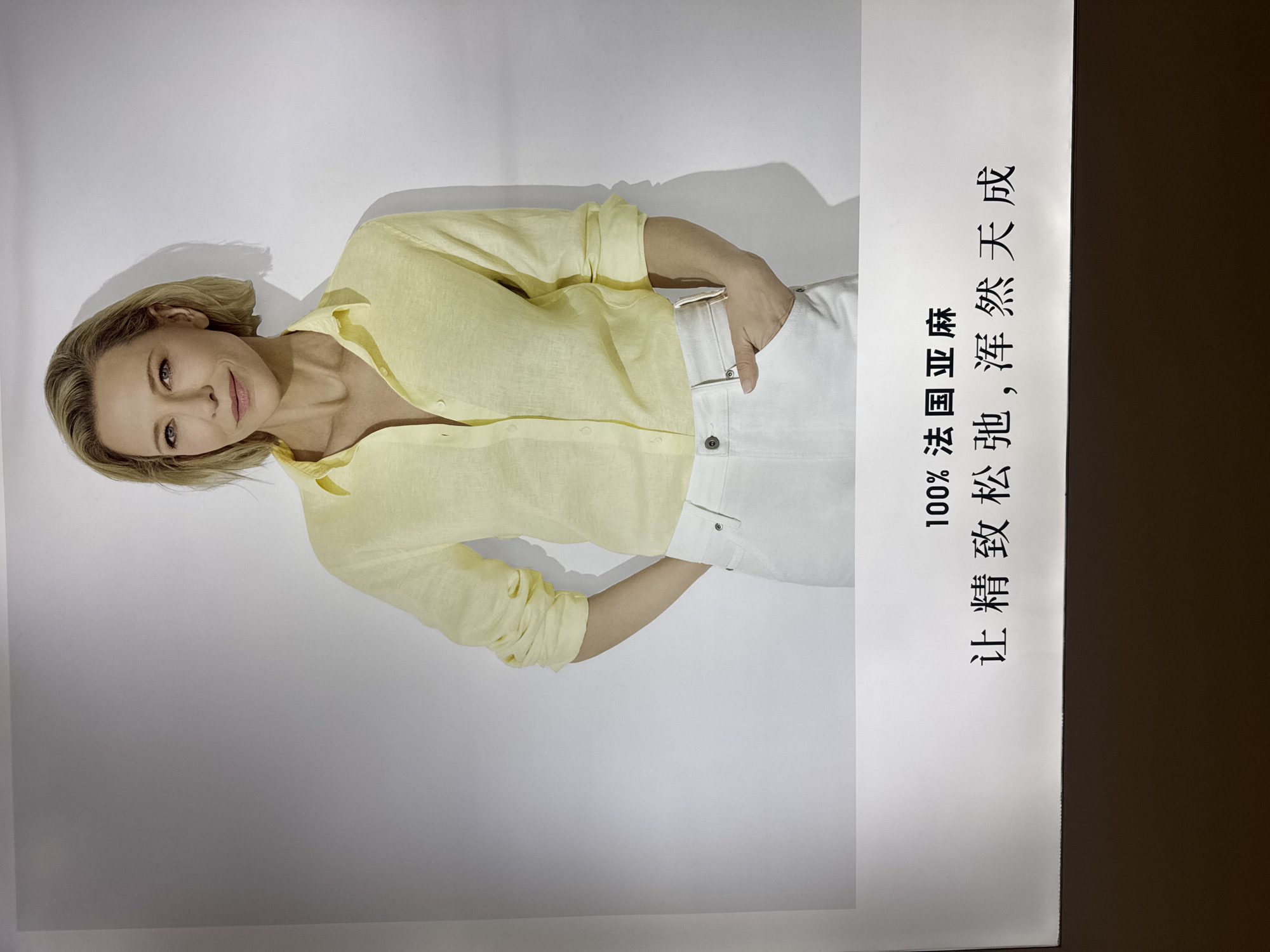 A medium shot displays Cate Blanchett, a woman with blonde hair, fair skin, and blue eyes, wearing a yellow linen button-down shirt and white pants. Her left hand is in her pocket. The background is a plain white wall with shadows. Below the image, Chinese text reads "100% 法国亚麻, 让精致松弛, 浑然天成". The image is presented against a dark brown background, likely a display in a public space, possibly in the Chaoyang District of China.