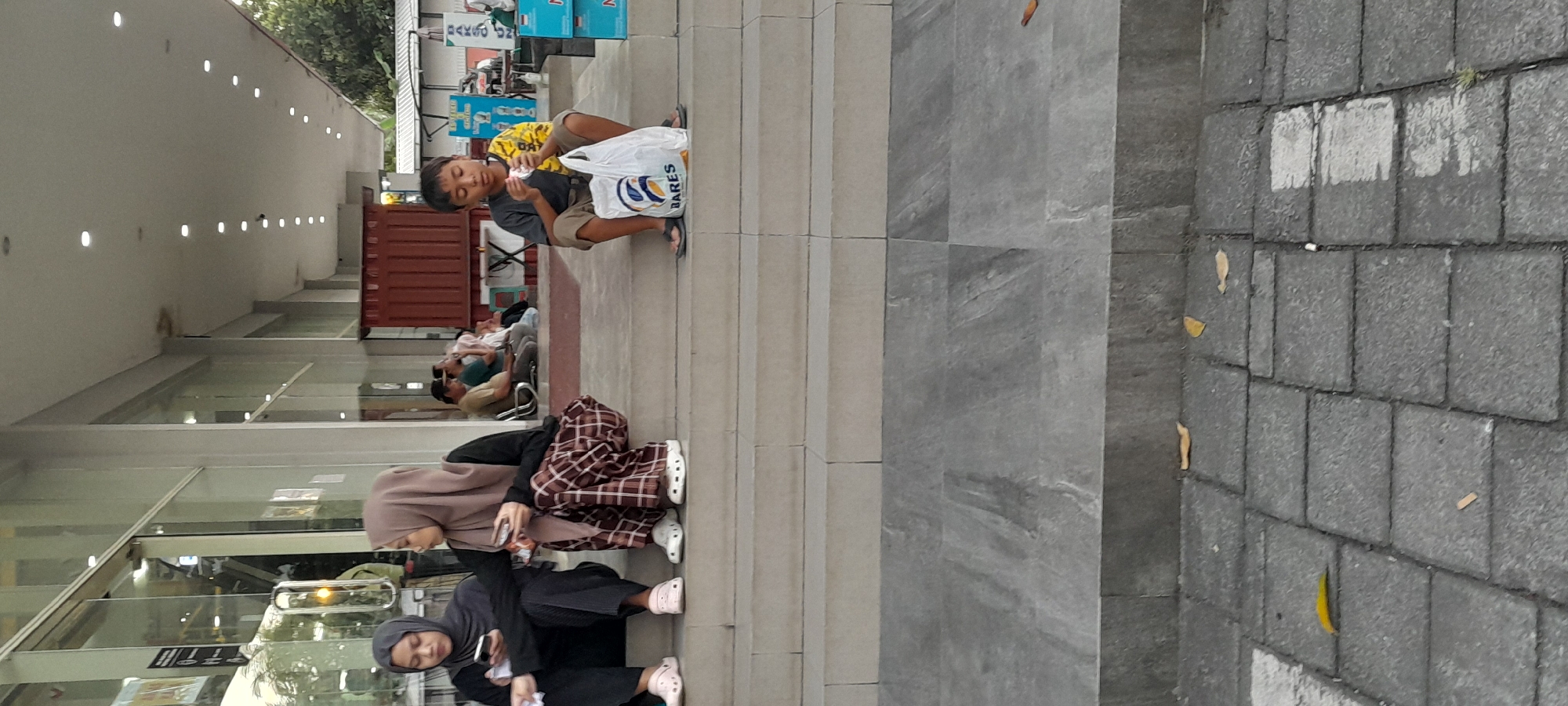 The image captures a scene outside a building with a covered walkway and outdoor seating area. In the foreground, two women are seated on steps, both wearing hijabs. The woman on the left is wearing a dark outfit and looking at her phone, while the woman on the right is wearing a patterned skirt and also appears to be holding a phone. To their right, a young boy is seated on the steps, holding a food item and a white shopping bag with the word "BARES" visible on it. Further in the background, other people are seated on benches and at tables. The environment suggests a public space, possibly a food court or commercial area, with grey tiled steps and a paved walkway in the foreground. The building behind appears to be a retail establishment with large glass windows. This scene is located in Rogojampi, Indonesia, a town in Banyuwangi Regency, East Java.