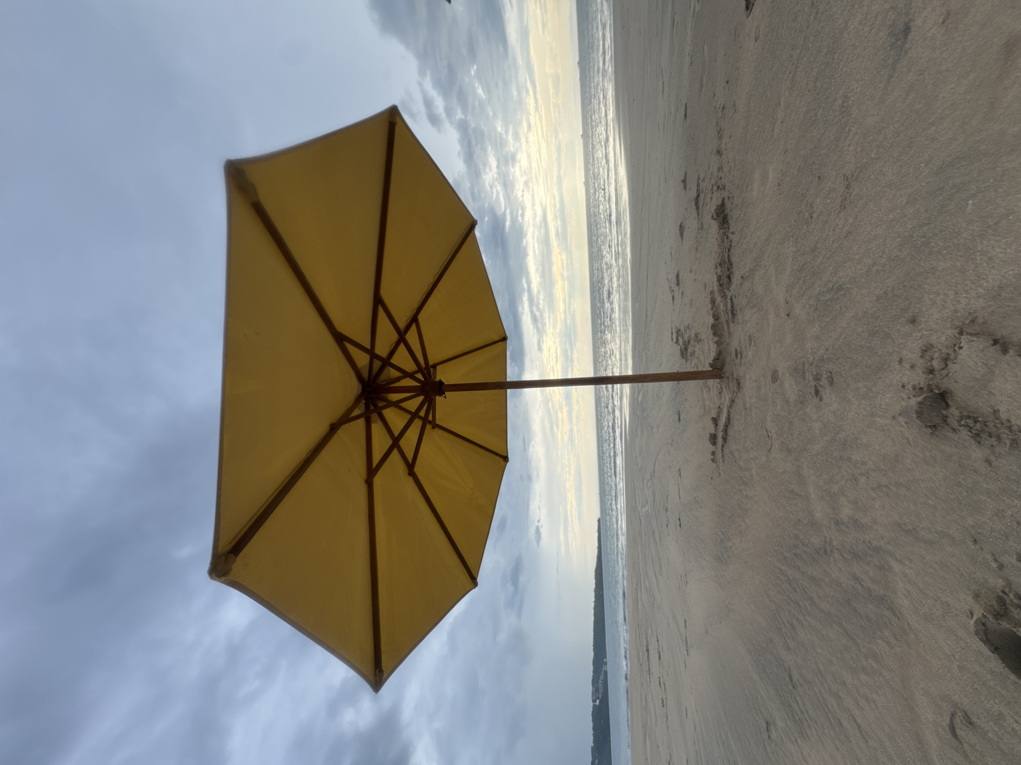 The image displays a serene beach scene, oriented vertically but depicting a horizontal landscape. A large, yellow beach umbrella with a visible wooden frame is prominently positioned on the left side, angled slightly upwards. Its canopy occupies a significant portion of the foreground and midground.
Beyond the umbrella, a vast, sandy beach extends towards the right, meeting the calm waters of the ocean. The sand shows subtle patterns and textures. The ocean surface appears relatively smooth with gentle ripples, stretching towards a distant horizon.
The sky is partly cloudy to overcast, with soft, diffused light suggesting either early morning or late afternoon. Lighter clouds near the horizon hint at a possible sunrise or sunset, casting a tranquil glow. No specific activity or event is taking place, but the setup implies a relaxing moment on the beach.
The setting is Kuta Selatan, Indonesia. There are no people visible, nor any discernible text. The overall atmosphere is peaceful and calm.