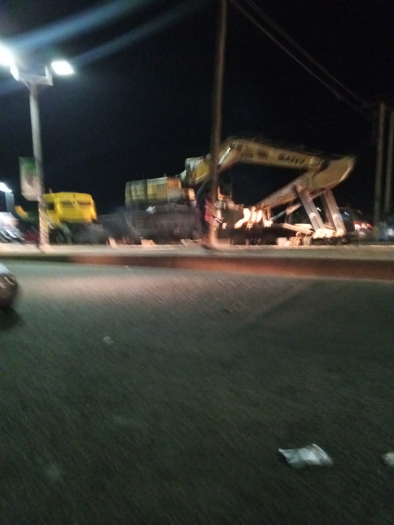 At night in what is possibly Bukuru, Nigeria, a yellow truck is parked alongside a large excavator being transported on a flatbed trailer. A street light illuminates the scene, and a person stands near the excavator. The foreground shows an asphalt road with a piece of litter.