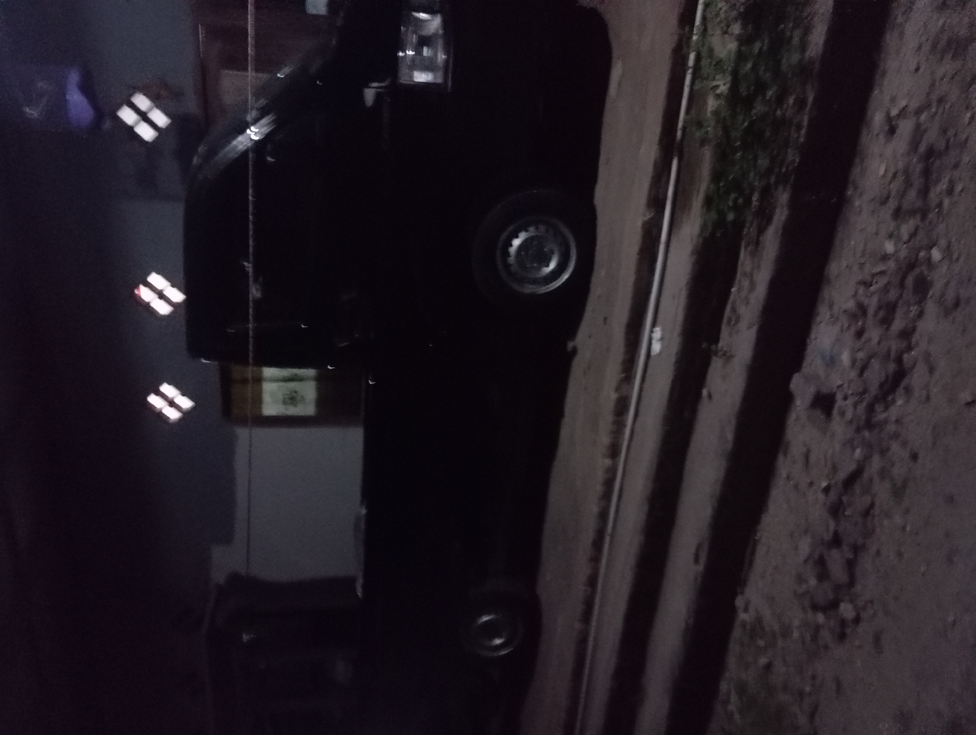 The image depicts a black pickup truck parked outdoors at night, possibly in a residential area of Rejosari, Indonesia. The truck is dark and silhouetted against the dim lighting, making precise details difficult to discern. Its headlights appear to be off, and there are reflections on its surface, suggesting it might be raining or the area is damp. The background shows a building with decorative square lights on its facade. The ground in front of the truck is paved and appears somewhat uneven, with some debris scattered around. A white pipe runs horizontally in the foreground, possibly indicating a drainage system or utility line. The overall scene is dark and atmospheric, with minimal visible activity or human presence.