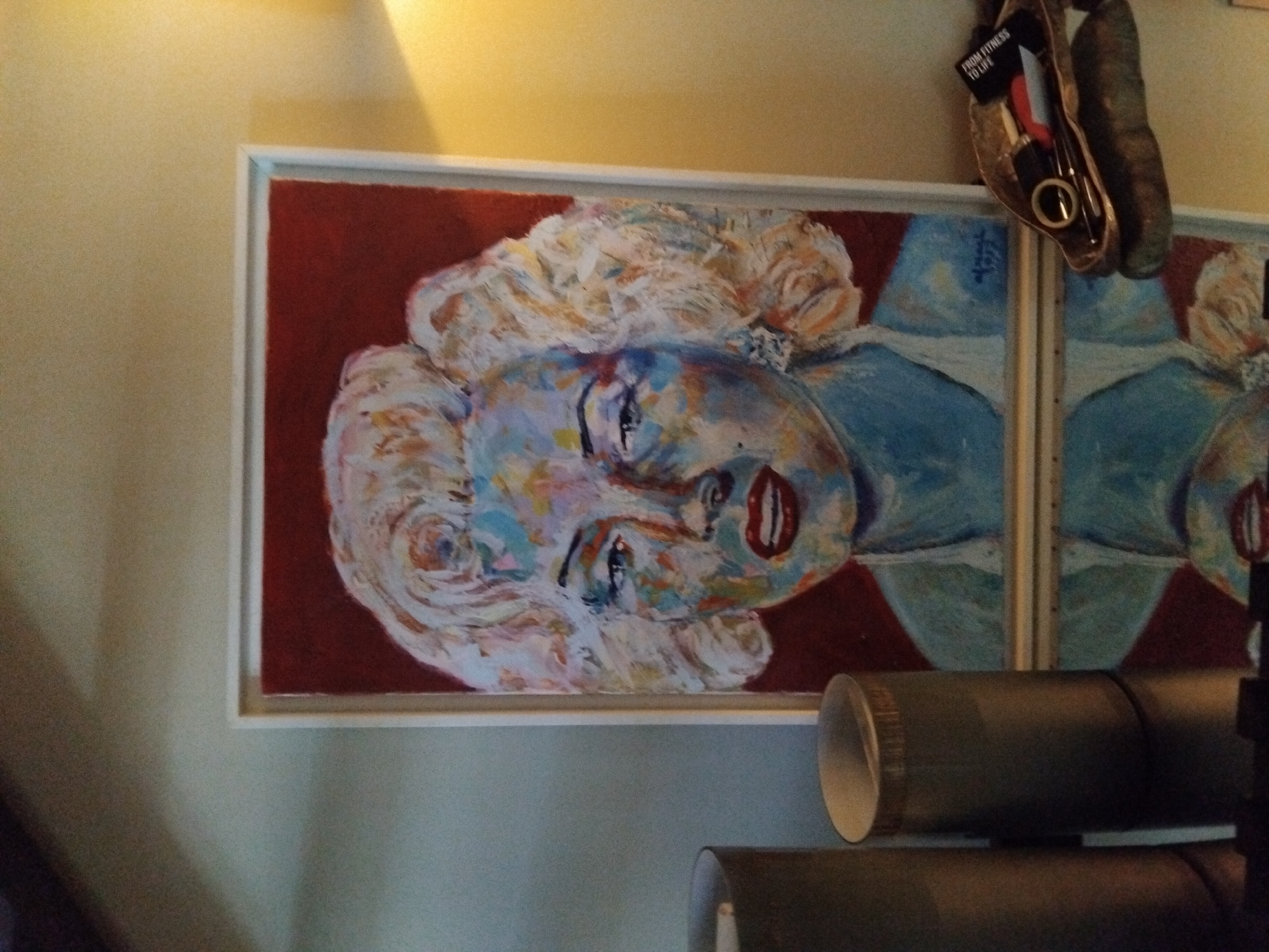 The image depicts a framed abstract portrait of Marilyn Monroe, rendered in vibrant colors with visible brushstrokes. The artwork is displayed on a wall with a subtle texture, illuminated by warm ambient light, suggesting an indoor setting. To the left of the portrait, two cylindrical objects, possibly vases or lampshades, are partially visible. Below the artwork, a reflective surface creates a mirrored image of the painting and the surrounding objects. In the lower right corner, a decorative tray holds several small items, including what appears to be a pair of scissors, a ring, and a small black tag with white lettering. The overall atmosphere is that of a home or a gallery space. No specific details indicate a location within Vila Nova de Famalicão, Portugal.