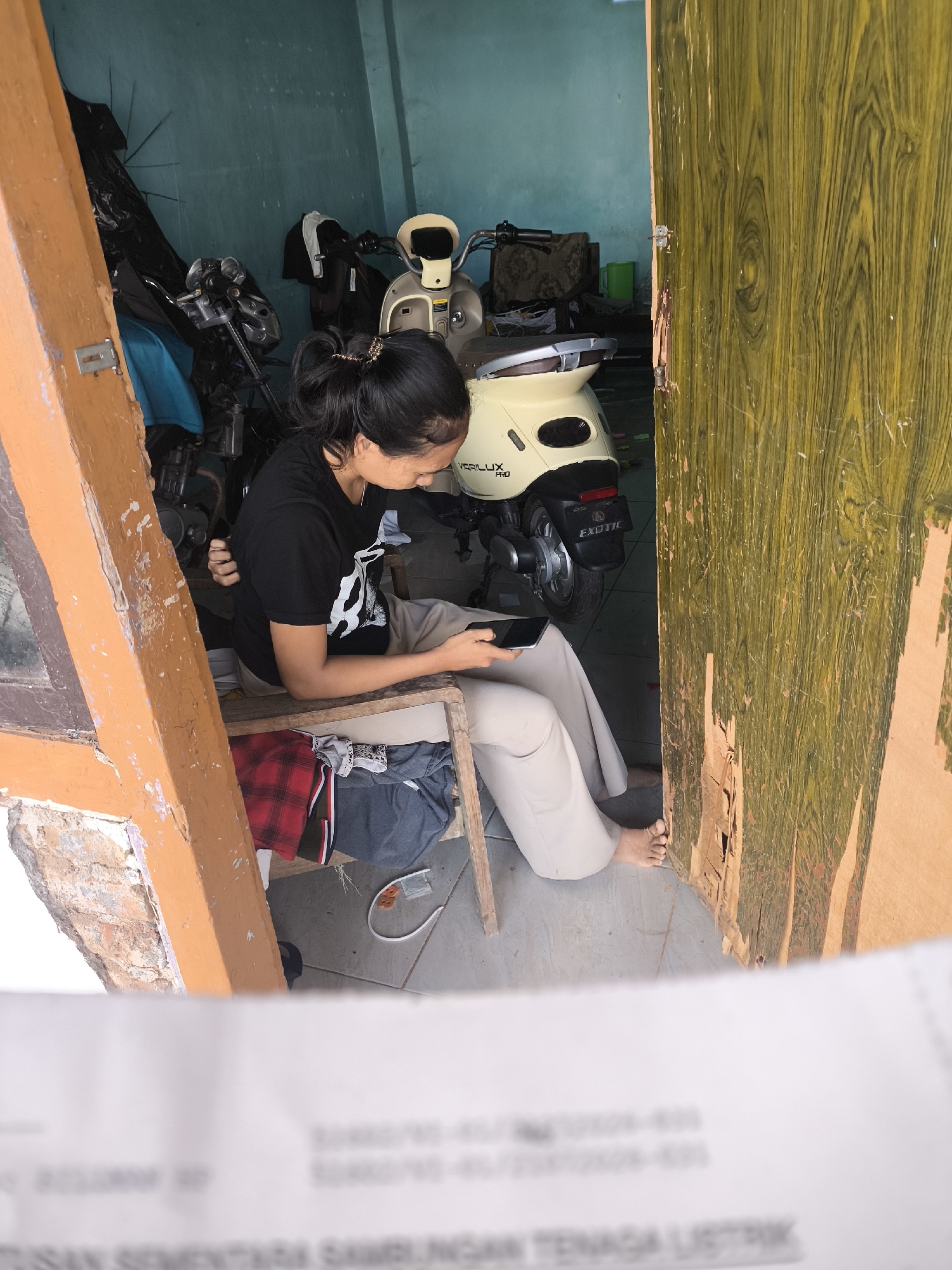In Lumajang, Indonesia, a young woman sits on a wooden chair, engrossed in her smartphone. She wears a black t-shirt and light-colored pants. The chair, positioned within an open doorway, has a pile of folded clothes beneath it. Behind her, two motorbikes are parked in a room with teal-colored walls. One motorbike is a cream-colored electric scooter, with "GARILLUX PRO" visible on its side, and "EXOTIC" on the rear fender. The other motorbike is partially obscured. The doorway opens to a wooden door with a distressed, weathered finish, revealing a glimpse of the exterior. A blurred document with text is visible in the foreground.