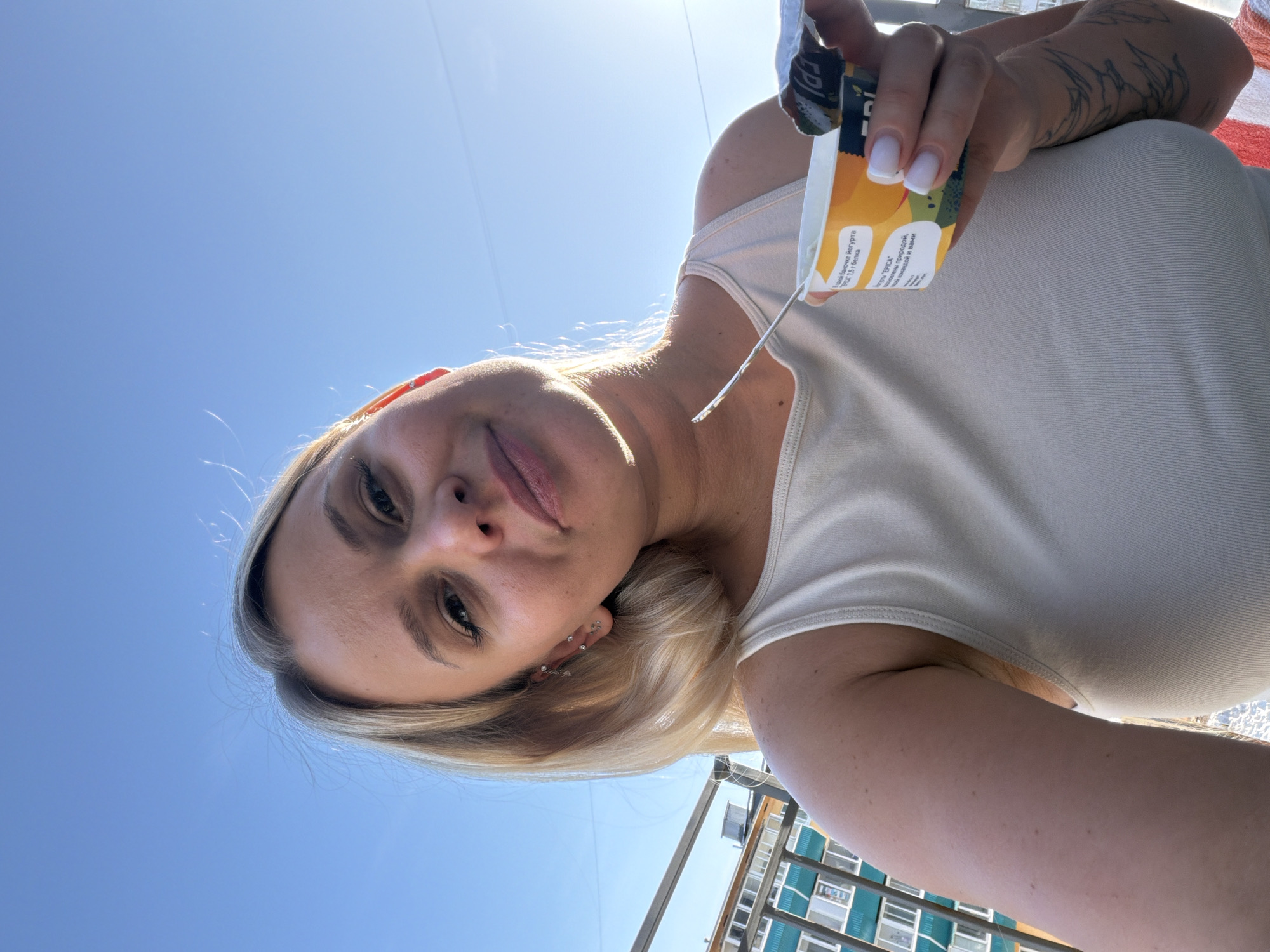 A woman is holding a yogurt container, facing the camera. She's blonde with multiple earrings in her left ear, wearing a white tank top. The sky is blue in the background. Part of a building with windows and railings is visible in the lower right corner. The yogurt container has the words "EPICA" on it and Russian text including "РАСА 1,5 г белка," "Едной баночке йогурта," and "кривлены природой." The location is not specified as Sarapul, Russia in the source image.