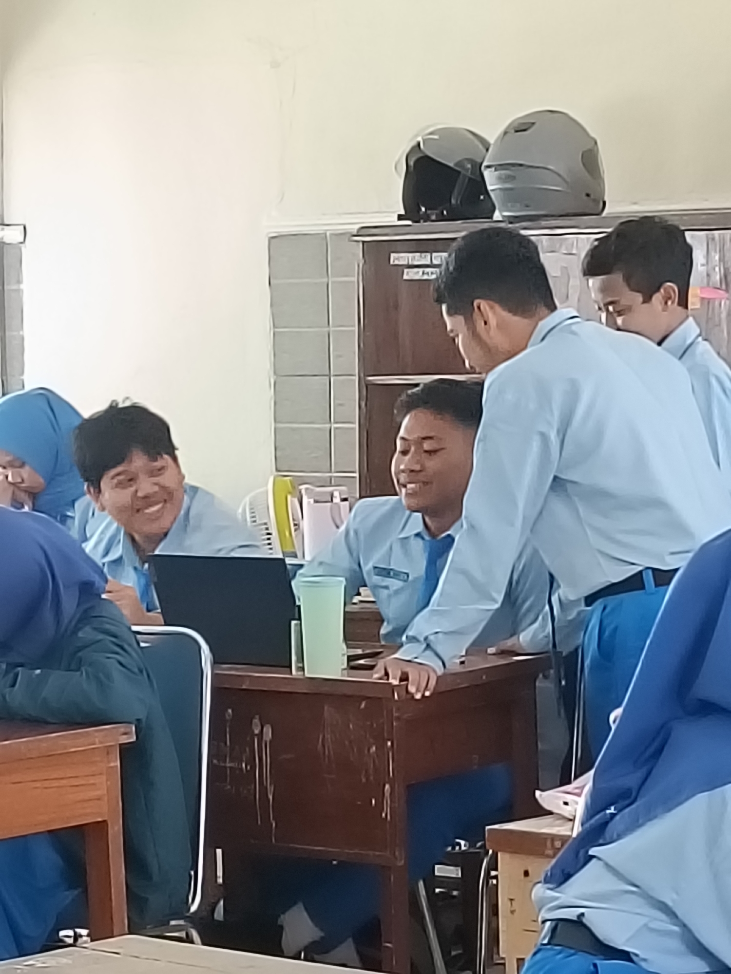 The image depicts a classroom setting in Kediri City, Indonesia. Several students, wearing light blue shirts and darker blue pants, are visible. One student is seated at a wooden desk with a laptop, smiling and looking to the left. Another student is standing behind this desk, leaning in and appearing to interact with the seated student. A third student is also standing behind the desk, further to the right. In the background, two helmets are placed on a shelf above a wooden cabinet. Other students and parts of the classroom environment, such as desks and chairs, are also present. The lighting suggests an indoor setting, likely during daylight hours.