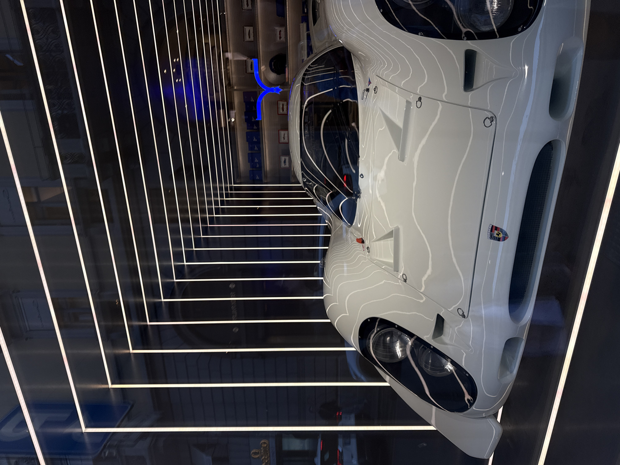 The photograph depicts a white sports car, likely a Porsche, displayed within a mirrored exhibit space. The exhibit's environment features parallel lines of light, creating a visually striking illusion of depth. Reflections in the mirrors add to the complexity and depth of the space. In the background, blurred objects and architectural details are visible, suggesting the location is a retail space. Given the context of a Porsche vehicle, the location is likely the Porsche store in Milan, Italy. No people are visible.