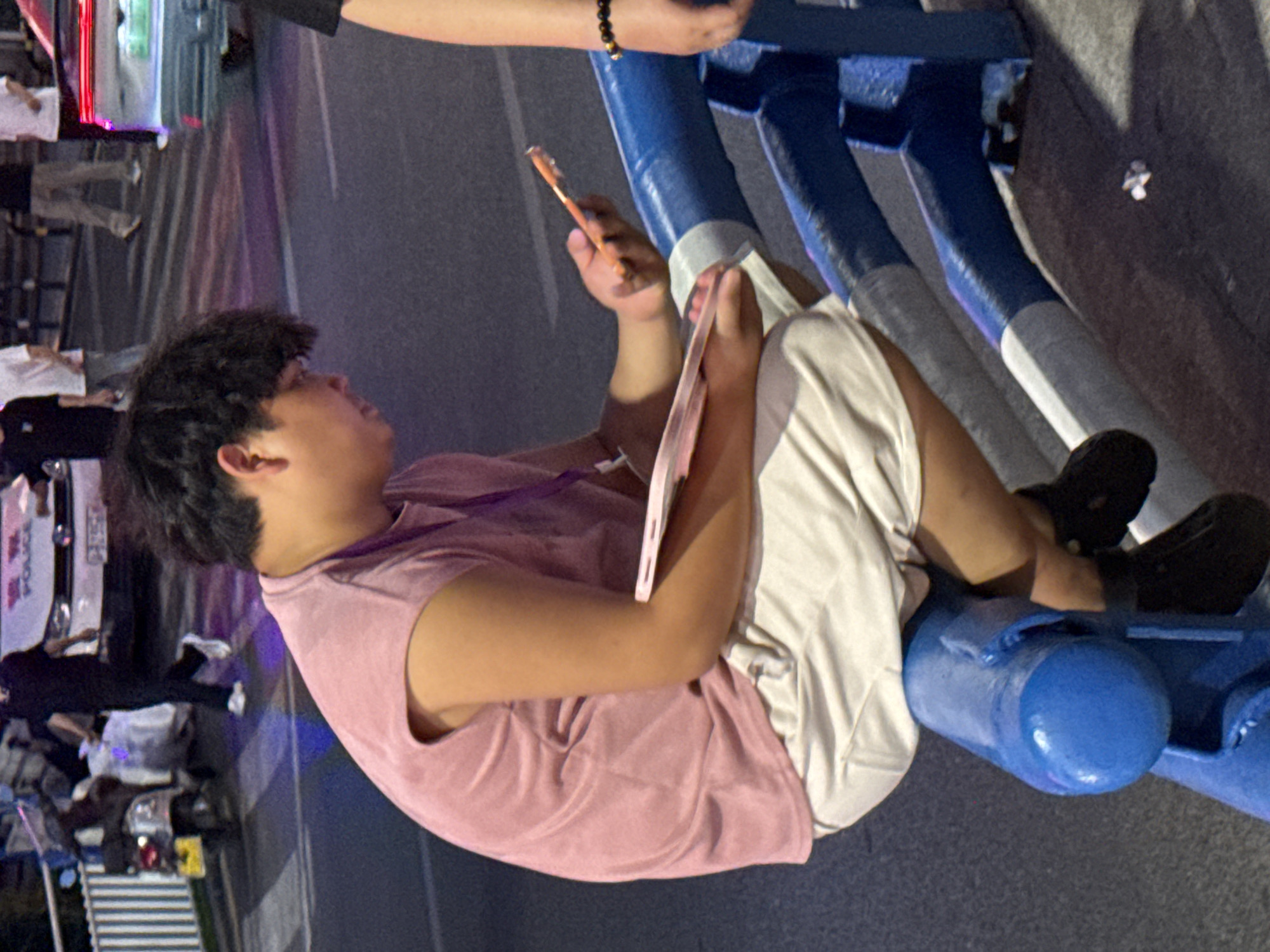 In Yuzhong District, China, a young East Asian male is seated on a blue and gray metal barrier. He is wearing a pink sleeveless t-shirt and light-colored shorts, with dark shoes on his feet. The individual is holding a pink object, possibly a phone or tablet, in his right hand. The background features a street scene with a white police car, a motorcycle, and several other individuals walking in the distance. The lighting suggests it is evening or night, with artificial lights casting colors on the road.