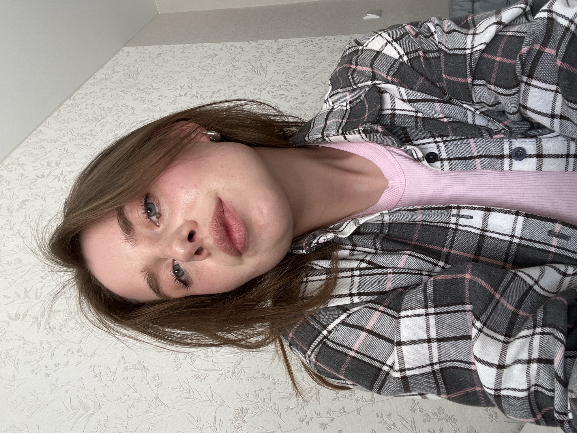 A young woman with light brown hair and blue eyes is centered in the image, wearing a pink ribbed top and a gray, white, and pink plaid flannel shirt. The background features a white wall with a subtle floral pattern. The location cannot be determined from the provided image.