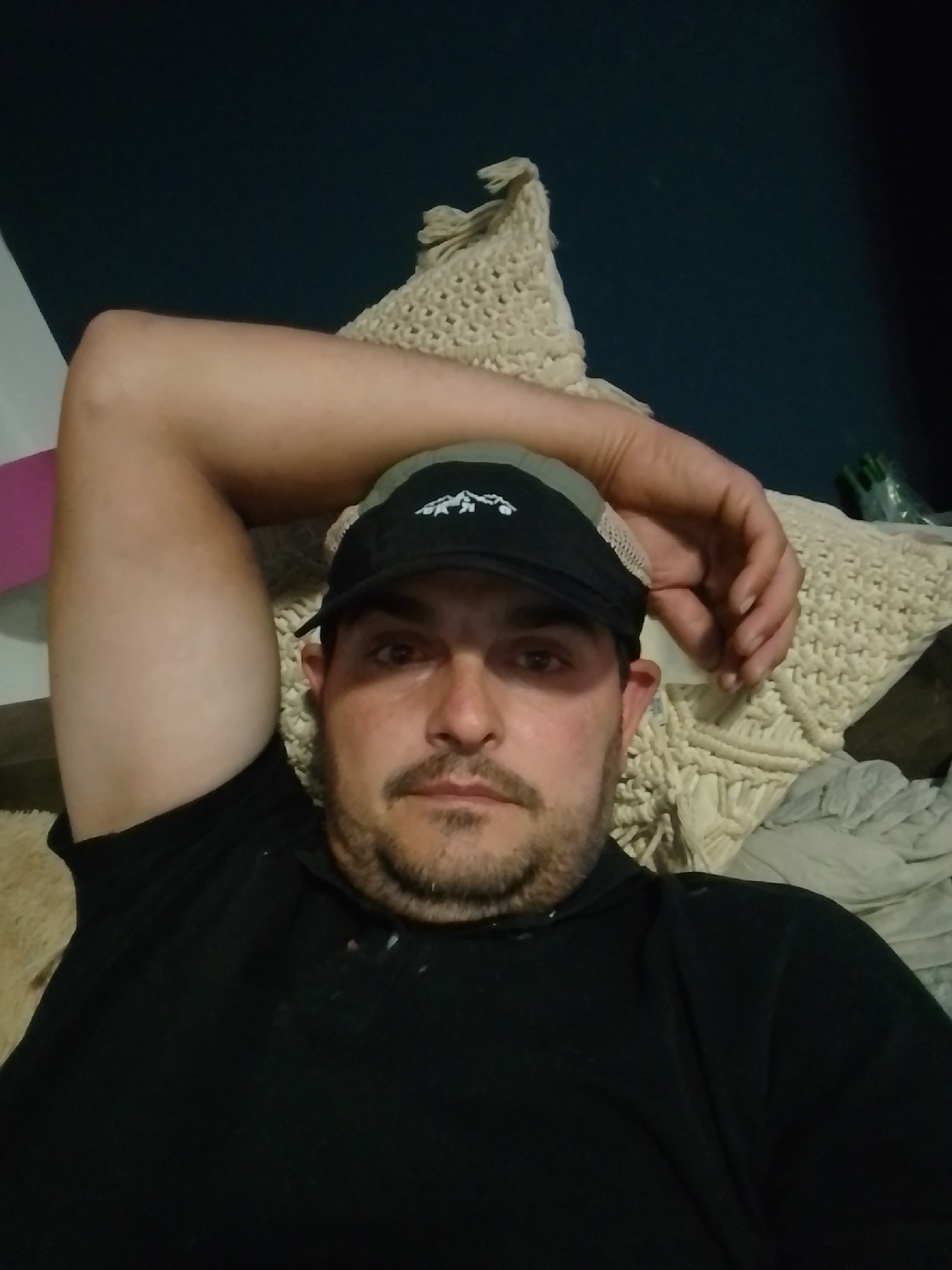 A man wearing a black t-shirt and a black baseball cap with a white embroidered logo is lying down, resting his head on his arm. His arm is bent at the elbow, and his hand is placed near his head. He has short, dark hair and a light brown beard and mustache. He is looking directly at the camera. The background consists of a dark blue wall and a cream-colored textured blanket or pillow. The lighting appears to be dim, suggesting it is nighttime or indoors with artificial lighting. There is no discernible location in the city of Wedel, Germany.