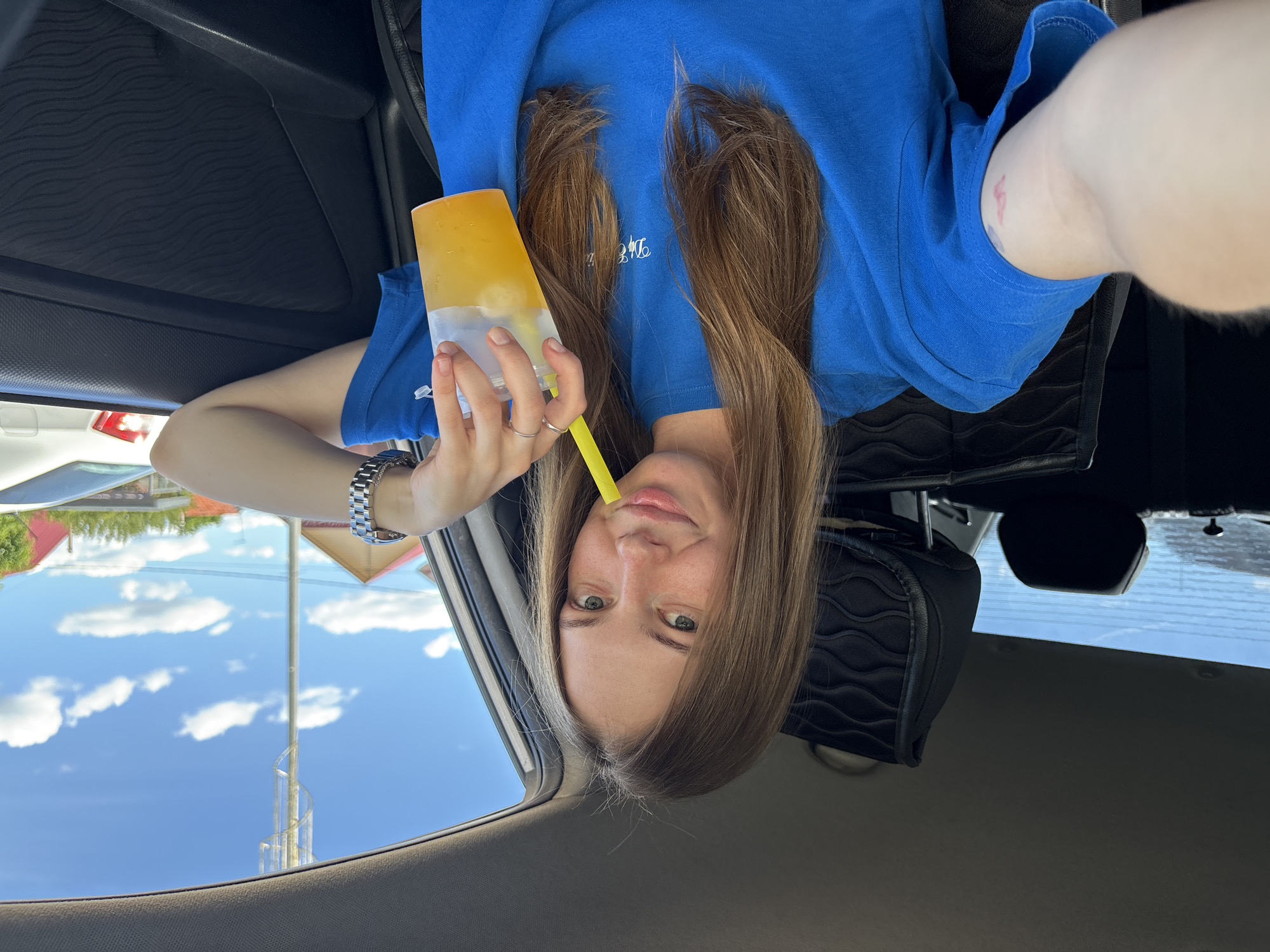 A young woman with long brown hair is sitting in the passenger seat of a car, holding a clear plastic cup with an orange beverage and a yellow straw. She is wearing a blue t-shirt and a watch on her left wrist. Her right arm is resting on the car door, and she is looking at the camera with a slight smile. Through the car window, a bright blue sky with white clouds is visible, along with houses and a cell tower. The interior of the car, including the headrest of the driver's seat, is visible. The image appears to have been taken on a sunny day. The location is the city of Perm, Russia.