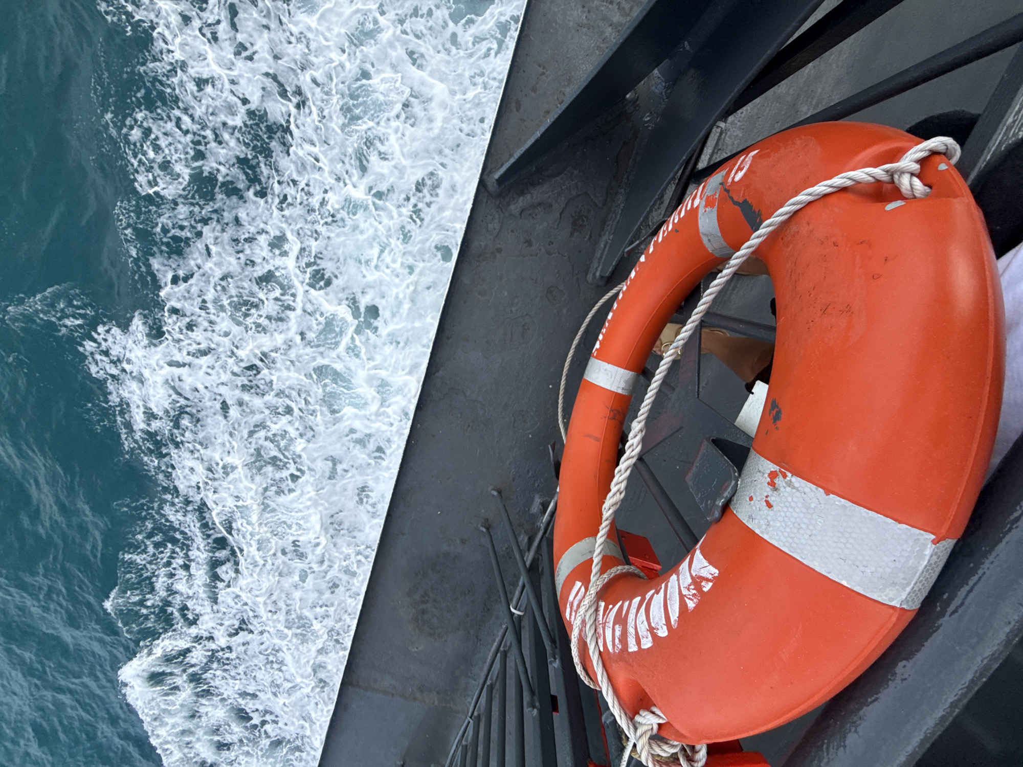 A high-angle, close-up shot features an orange life preserver with a white rope tied around it, resting on the dark, weathered deck of a vessel. The life preserver is positioned in the foreground, with its bright orange color contrasting against the dark background. White reflective tape is adhered in two bands around the life preserver, showing some wear and tear. The rope is secured with a knot. Behind the life preserver, the dark metal railing of the boat is visible. The background is filled with the dynamic movement of turquoise water, with white frothy waves and foam indicating the vessel is in motion. The scene appears to be set on a body of water, possibly the ocean, given the texture and color of the water. The specific location in Ko Samui District, Thailand cannot be determined from the image itself. There are no people visible in the frame.