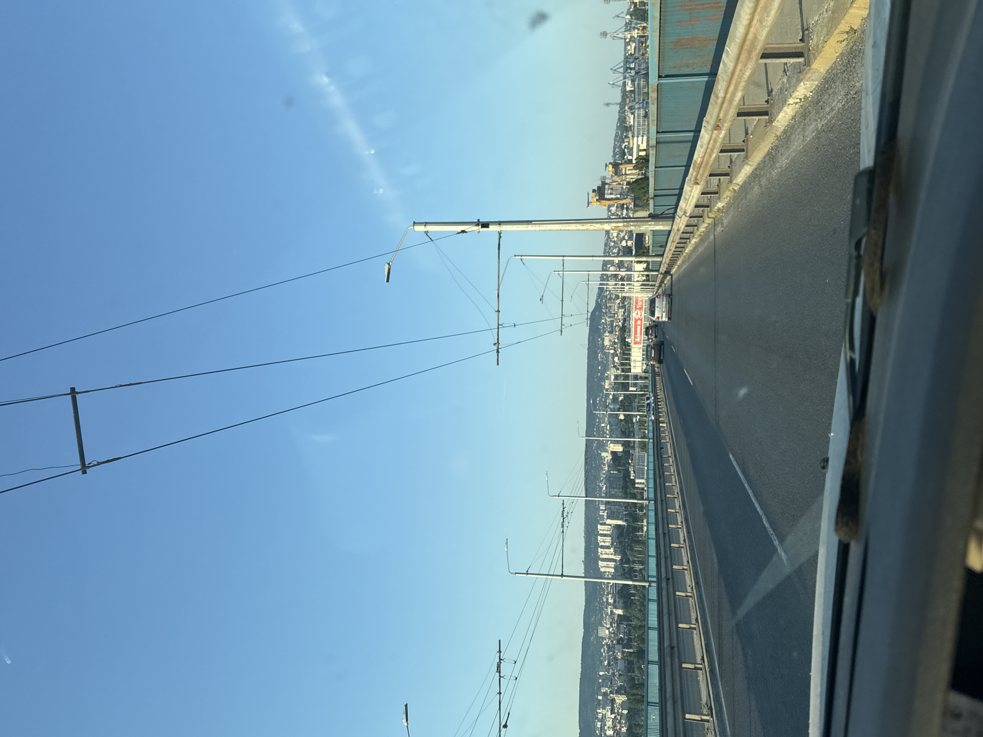The image depicts a view from a vehicle driving on a bridge over a river, overlooking a city skyline. The sky is clear and blue. Overhead wires, likely for trolleybuses, crisscross the frame. Streetlights are visible along the bridge. In the distance, a dense urban area with numerous buildings stretches across the landscape. On the right side of the bridge, a blue metal railing with rusted support posts runs along the edge. The road ahead is paved and has lane markings. Several cars are driving on the bridge, heading in the same direction as the viewer. The overall impression is that of a daytime commute in an urban environment.