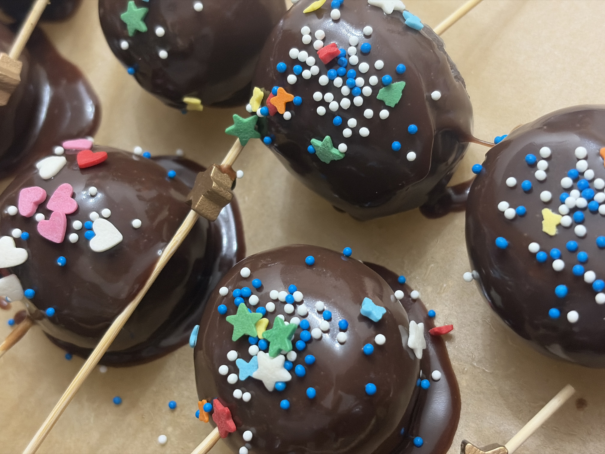 The image shows a close-up of several chocolate-covered cake pops. Each cake pop is covered in a dark chocolate coating and decorated with colorful sprinkles of various shapes, including hearts, stars, and small round candies in white and blue. The cake pops are skewered with wooden sticks and rest on a light brown surface. There is no indication of Saint Petersburg, Russia, in the image.