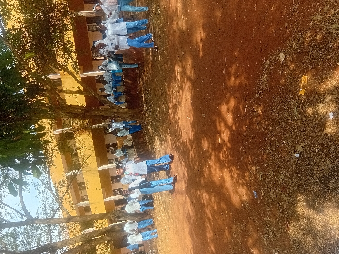 A group of people stand in front of a yellow building, possibly a school or institution. The individuals appear to be students, clad in light-colored shirts and blue pants. A tree with dense foliage frames the left side of the image. The ground is predominantly brown and earthy, with scattered debris visible. The picture's orientation is vertical.