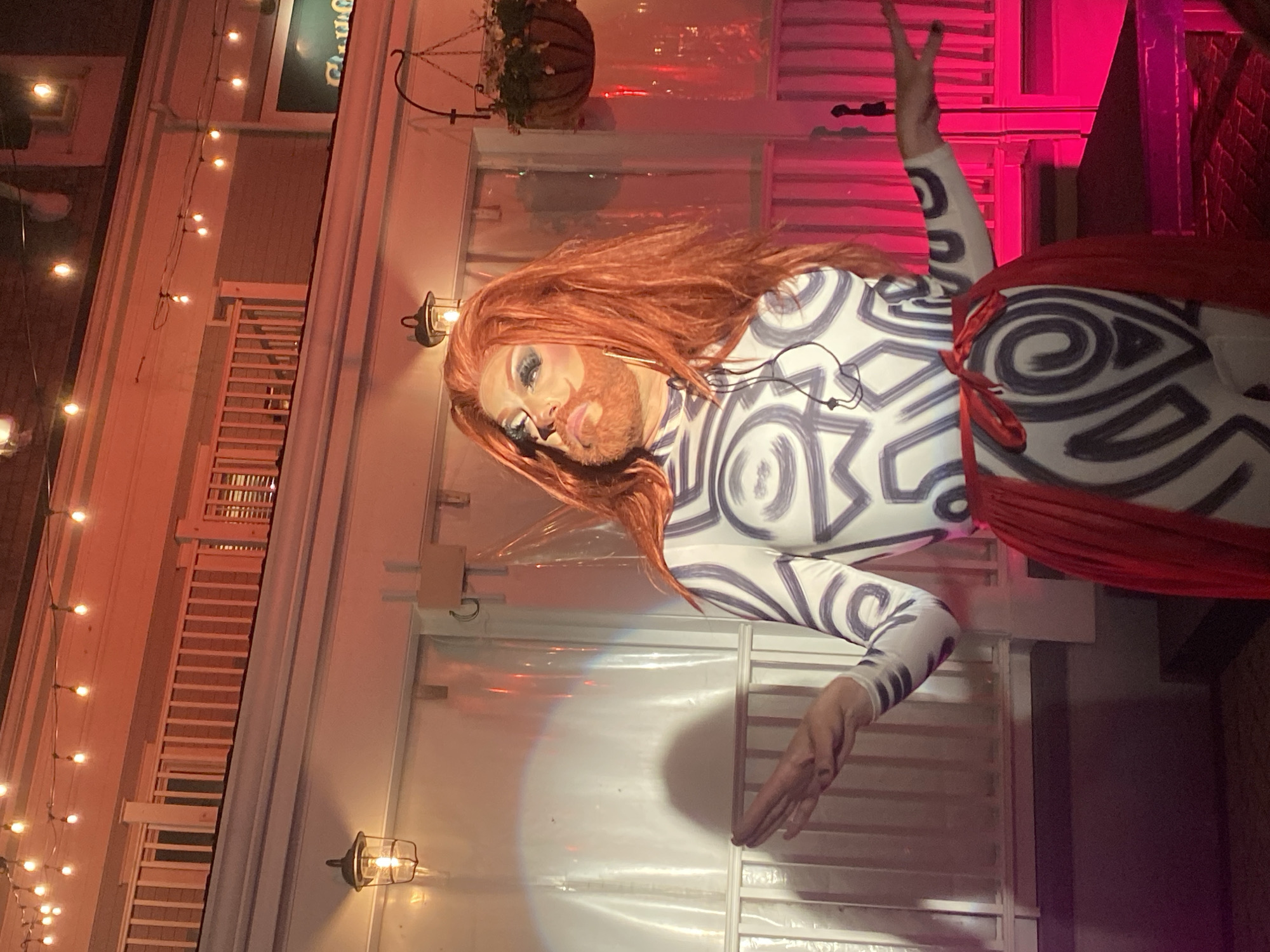 In Provincetown, Massachusetts, a drag performer with vibrant red hair and a full beard stands on a stage outdoors, illuminated by pink lights. The performer is wearing a white, full-body suit with a bold, abstract blue pattern and a bright red sash tied around their waist. Their makeup is dramatic, with dark eyeliner and vibrant eyeshadow. Behind the performer, a building with string lights is visible, with a balcony above. A hanging basket filled with flowers adds a touch of nature to the scene. The overall atmosphere is festive and theatrical.