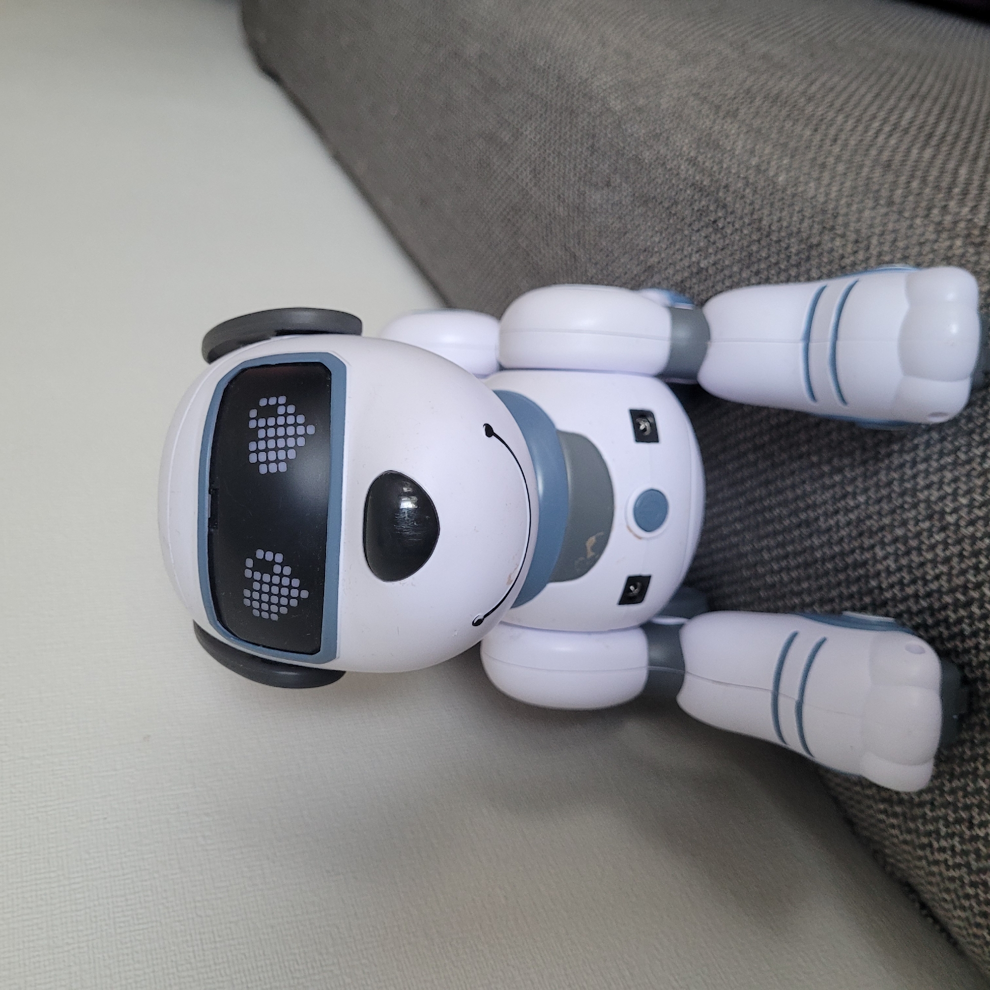 A white robot dog with gray accents sits on a gray upholstered couch against a lightly textured off-white wall. The robot has a black nose and a smiling mouth drawn on its face, and its eyes are displays showing dotted heart shapes. Its ears are gray and rounded, and its body is white with blue trim. The robot has four legs, each ending in a paw-like foot. The background wall is lightly textured and plain. The couch is upholstered in a dark gray fabric with a subtle woven pattern. The lighting in the scene is soft and diffused, suggesting an indoor environment. There are no people or other animals present in the image. The robot is positioned in the center of the frame, facing forward. The image appears to be a close-up shot, focusing on the robot. The location is Jūrmala, Latvia.