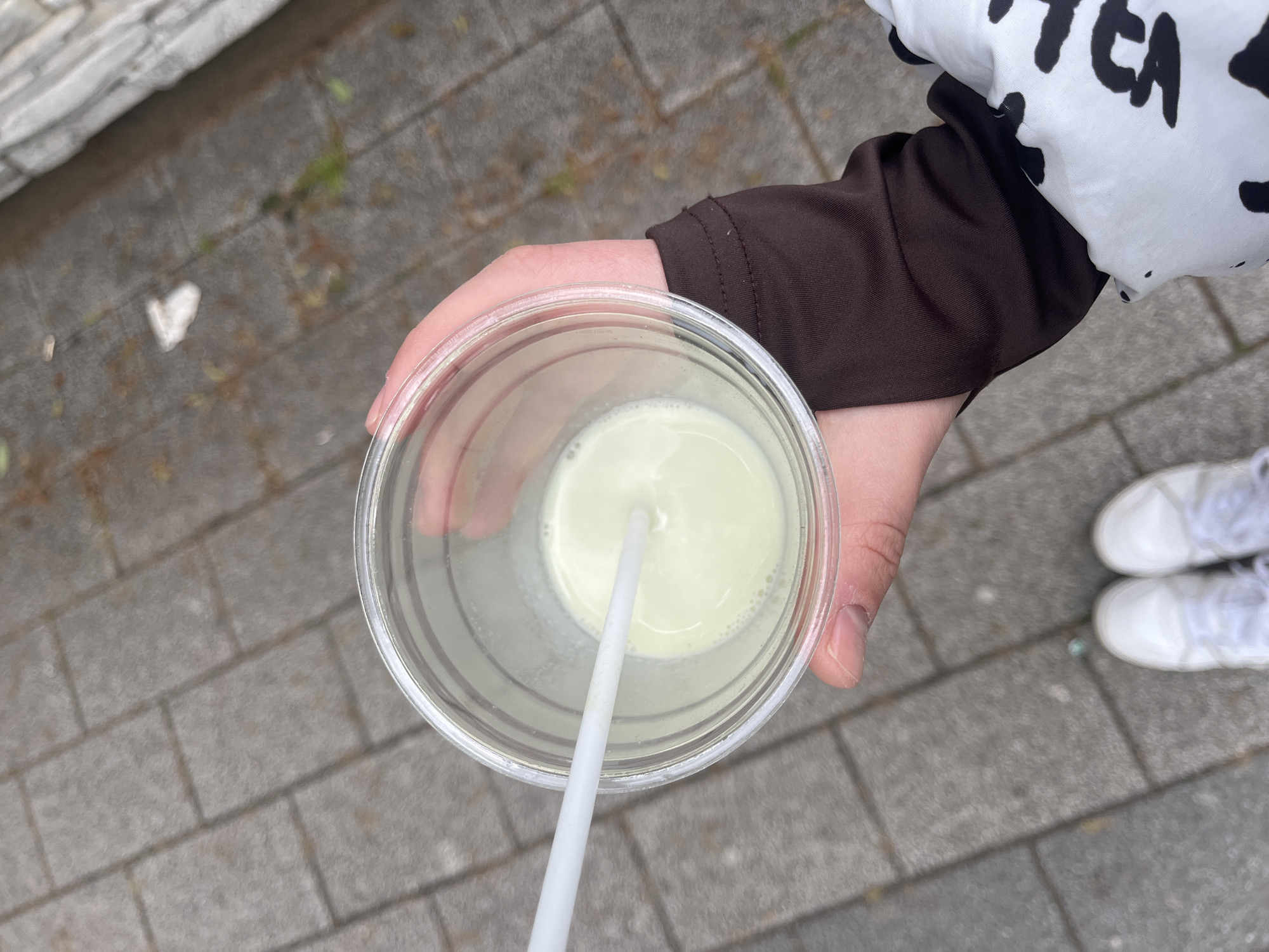 A person holds a clear plastic cup with a light green liquid and a white straw. The person wears a dark brown long-sleeved shirt under a white and black patterned jacket. The background shows a gray brick walkway. A pair of white shoes is partially visible in the lower right corner. The image does not provide enough information to determine if the location is in Plovdiv, Bulgaria.