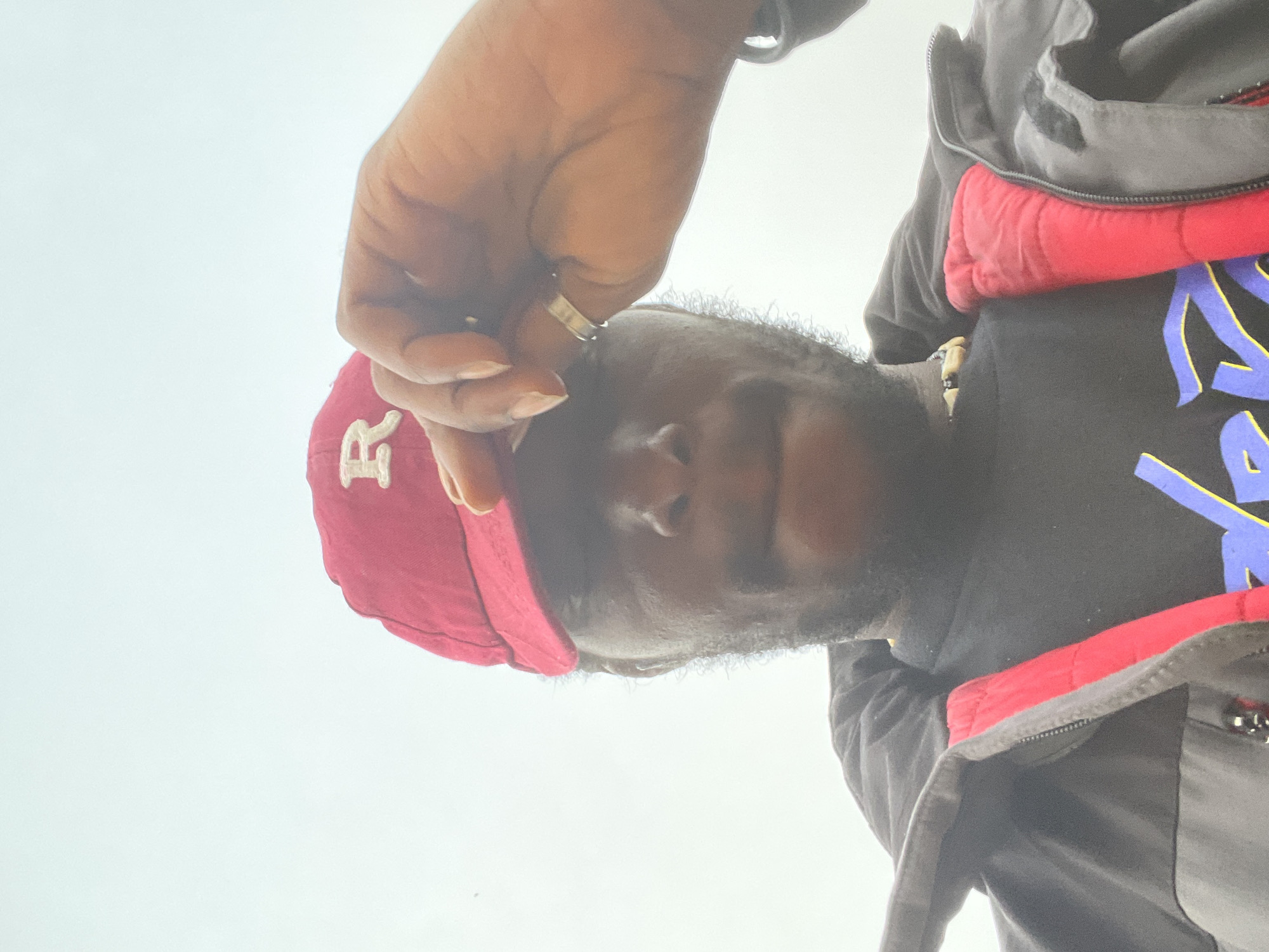 A man is shown from a low angle, appearing to look down at the camera. He is wearing a red baseball cap with a white "R" on the front, a black T-shirt with a colorful design, and a jacket with red trim. He has a beard and is wearing a ring on his hand, which he is using to touch the brim of his cap. The background is a pale sky. There is no clear indication that the location is Jos, Nigeria.