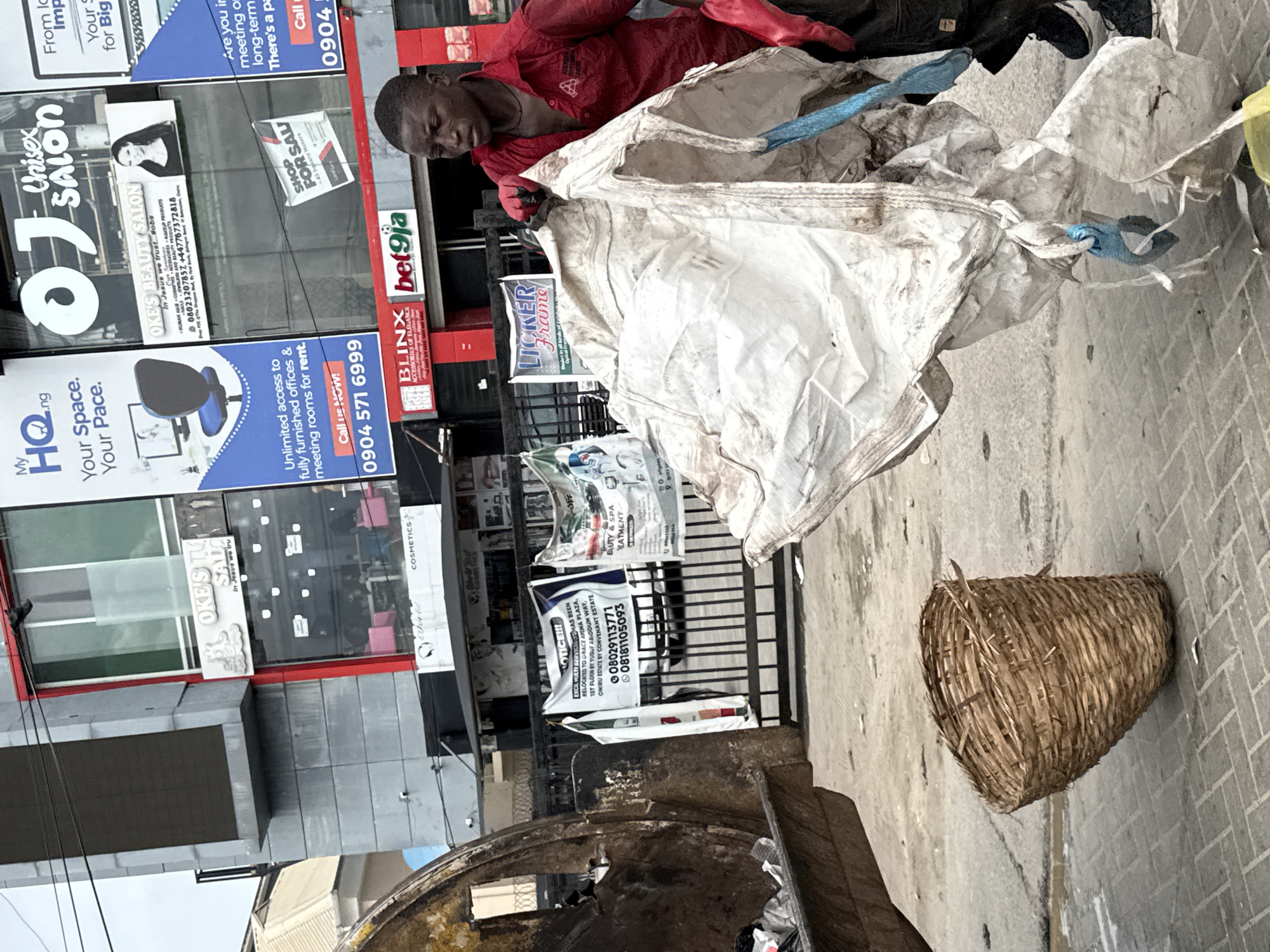 The image shows a man in Ilado, Nigeria, likely a sanitation worker, standing over a metal trash receptacle. He is wearing a red shirt and red gloves and is in the process of emptying a large, dirty white sack into the container. Beside the receptacle, on a paved surface, rests a woven basket. The background features several commercial buildings covered in advertisements for services such as real estate rental, beauty salons, and cosmetic products. The OCR text on the signs includes phone numbers beginning with 0904 and 0802, suggesting the location within Nigeria.