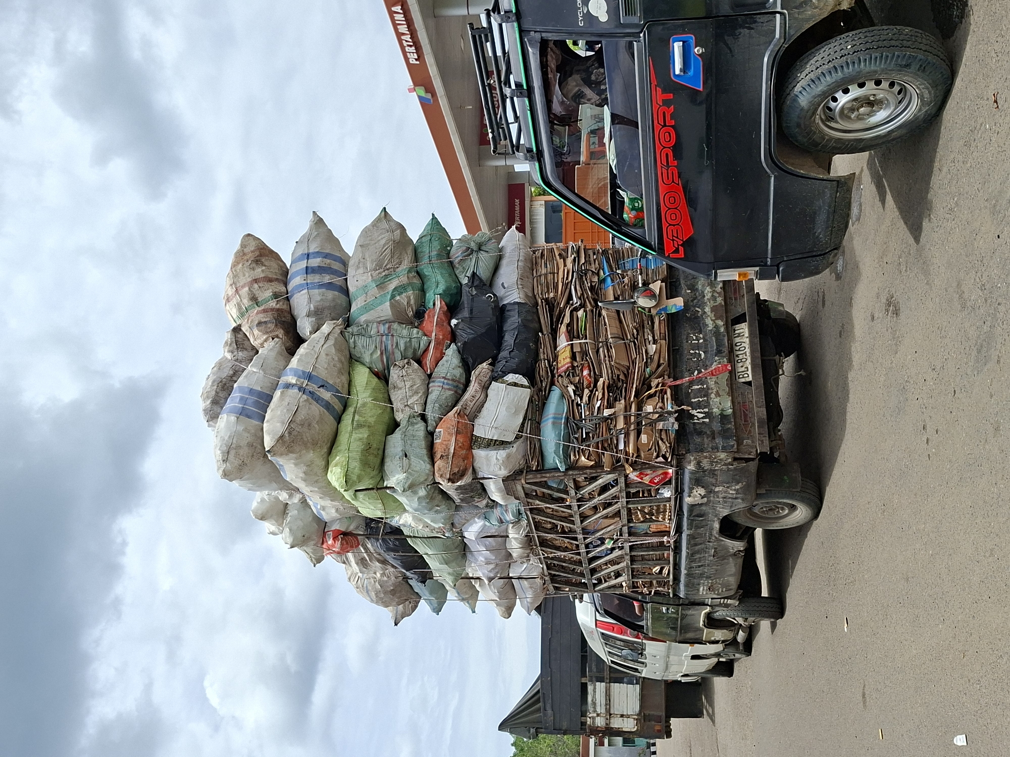 A truck overloaded with bags and cardboard is parked near a Pertamina gas station in Lhokseumawe, Indonesia. The truck, a black L300 Sport, has a visible license plate reading "BL 8169 NT". The bags, various colors and patterns, are stacked high above the truck bed. The surrounding environment shows an overcast sky and the gas station's infrastructure.