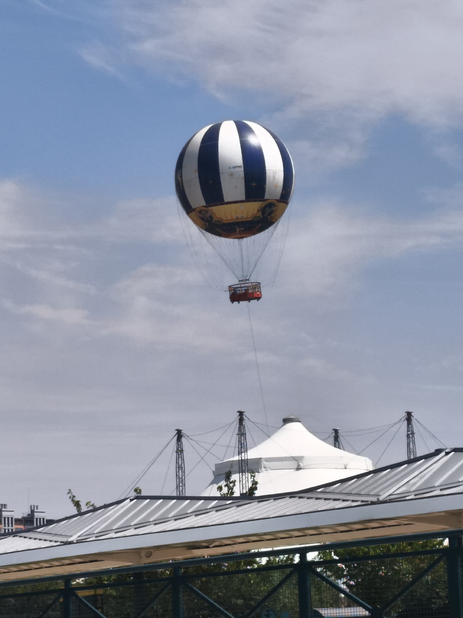 A hot air balloon with white and dark blue stripes and a yellow and black lower section is flying in a light blue sky with scattered clouds. Below the balloon, suspended by thin cables, is a small, red gondola. In the background, a large white tent-like structure with supporting poles and cables is visible, along with a portion of a modern building with a sloped, metallic roof. The scene appears to be set in an outdoor entertainment venue, possibly related to Disneyland Paris in Chessy, France, given the distinctive architecture and the presence of a tethered balloon ride often found in theme parks. The overall impression is of a bright, clear day with recreational activity taking place.