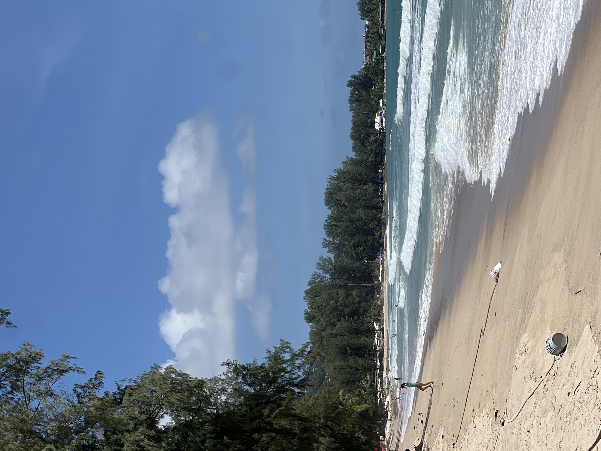 The image depicts a wide, sandy beach on a sunny day in Choeng Thale, Thailand. The foreground is dominated by sand, with a few pieces of debris including a white plastic bucket with blue lettering lying on its side, and a tangled rope extending diagonally across the sand. To the left, a person in a green swimsuit is walking away from the camera, towards the water. The middle ground shows the turquoise ocean with multiple rows of white, breaking waves. Further in the background, a line of lush green trees runs along the coastline, with scattered buildings and umbrellas visible amongst the foliage. The sky is a bright blue with scattered white clouds. Several other people are visible in the water and on the beach in the distance.