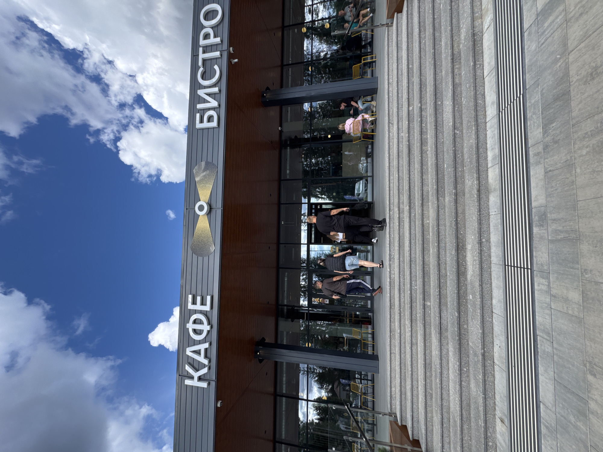 The image shows the exterior of a cafe and bistro. The building has a modern facade with large glass windows and a prominent sign that reads "КАФЕ" (Cafe) on the left and "БИСТРО" (Bistro) on the right, separated by a graphic of a propeller. A group of people are seen walking up a wide set of stone stairs leading to the entrance of the establishment. Inside the cafe, visible through the windows, are patrons seated at tables, some of whom are dining or conversing. The environment is outdoors, with a bright blue sky and white clouds overhead, suggesting a daytime scene. The architecture and signage indicate this is likely a commercial establishment in a developed area, possibly a suburban or recreational zone. The presence of trees in the background suggests it might be located near a wooded area. The image does not explicitly contain information that can place it in Lakhdenpokhya, Russia, though the Cyrillic signage is consistent with a Russian-speaking location.