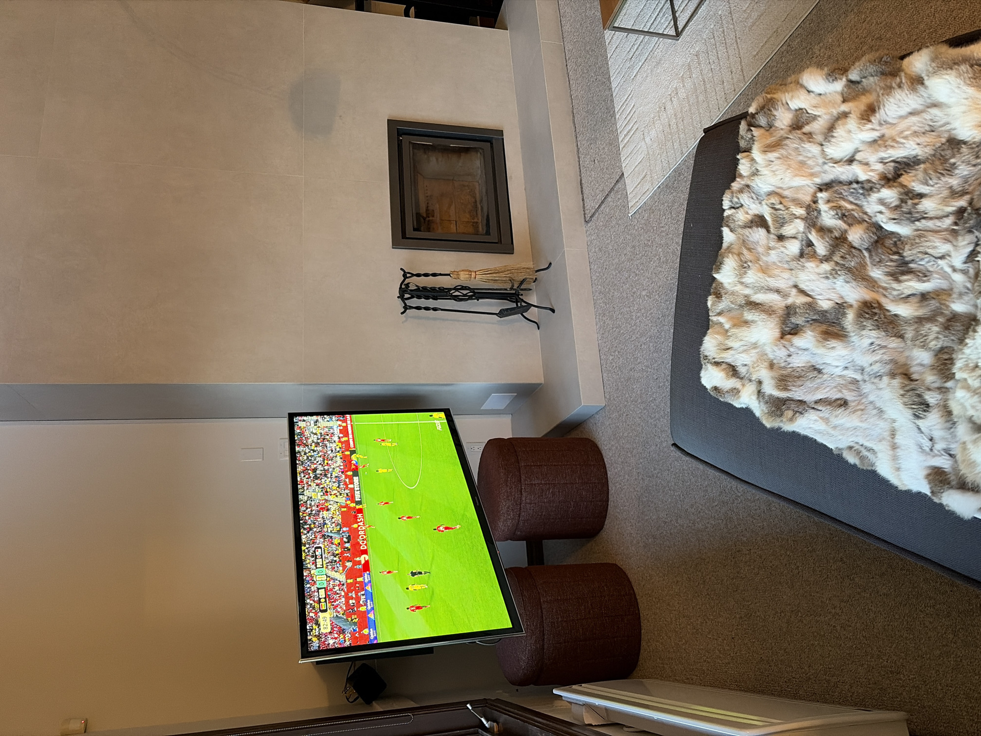 The image depicts an interior scene, likely a living room, possibly located in Mont-Tremblant, Canada, given the context. A large flat-screen television is mounted on the wall, displaying a soccer match. Below the television are two dark reddish-brown cylindrical ottomans. To the right of the television is a modern fireplace with a dark metal frame, set into a light-colored tiled wall. Fireplace tools, including a poker, tongs, and a brush, are neatly arranged on a stand next to the fireplace. A section of a grey couch is visible in the foreground, draped with a plush, multi-colored faux fur blanket. The flooring is carpeted. The environment appears to be a contemporary living space designed for relaxation and entertainment.