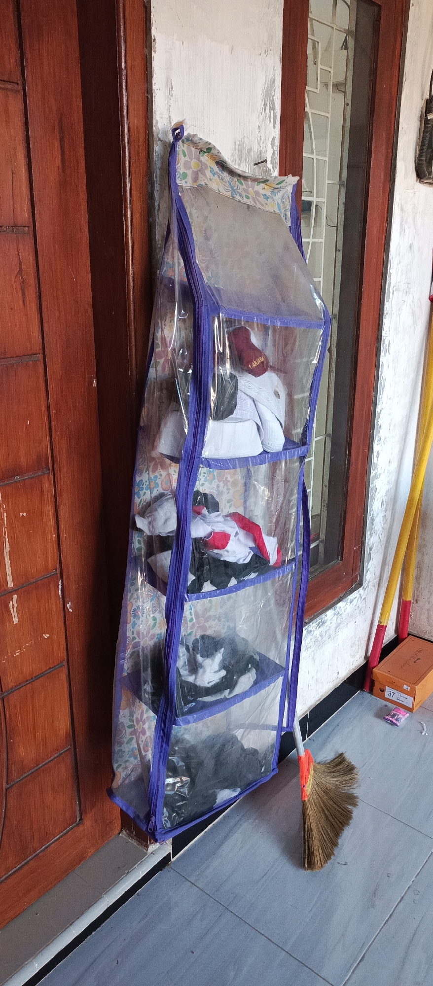 A floor-level, eye-level, full shot captures an outdoor scene. To the left is a dark brown, wooden door with vertical panels. To the right of the door is a clear plastic hanging organizer filled with various clothing items, primarily white and red. The organizer has purple trim and is attached to the wall. The wall itself is white and appears weathered. A window with a white lattice frame is visible behind the organizer. In the foreground, a gray tiled floor is visible, with a broom leaning against the organizer on the right. Two yellow poles with red ends stand behind the organizer. A small orange box with "37" printed on it sits on the floor to the right of the broom.