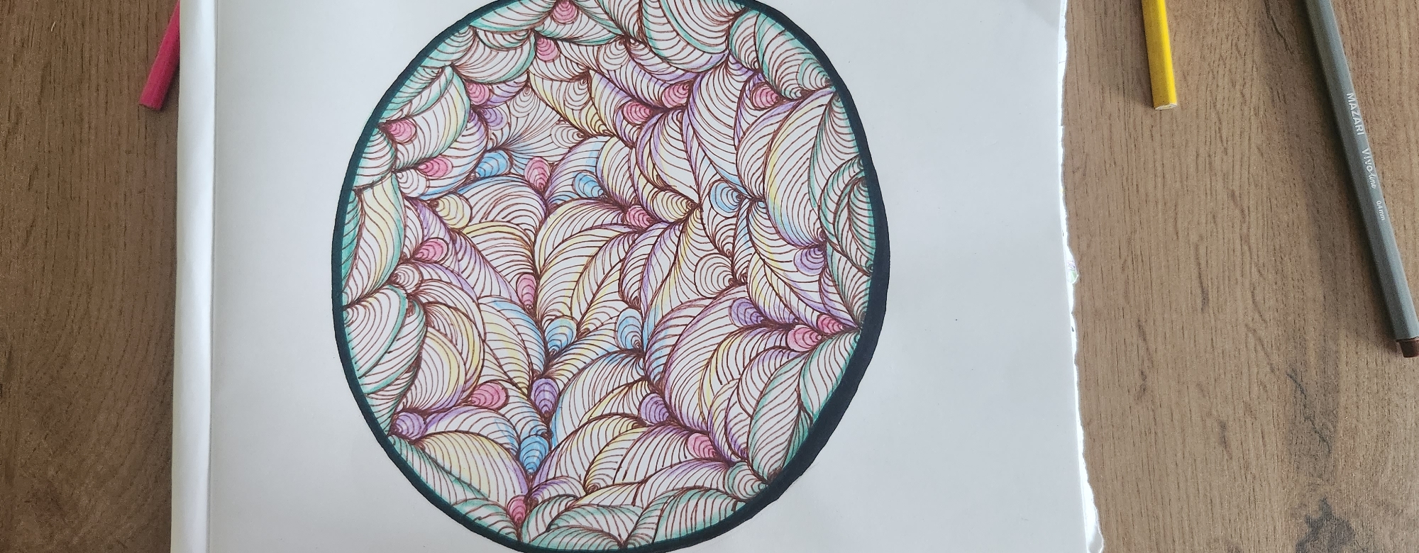 The image shows an oval-shaped drawing filled with a complex pattern of repeating, curved lines and small, teardrop-shaped elements. The lines are drawn in dark brown, and the teardrop shapes are colored with pastel shades of pink, yellow, light blue, and light green. The drawing is enclosed by a thick, dark green border. The drawing is on a white sheet of paper, which is placed on a wooden surface. Two colored pencils, one pink and one yellow, are visible on the wooden surface, as well as a gray pen with "MAZARI Vivo line 0.4 mm" printed on it. The upper part of the image shows a white border above the drawing, and a wooden surface beyond that. The lower part of the image shows the wooden surface extending downwards. The location in the city of Kazan, Russia is not depicted or suggested by the image.