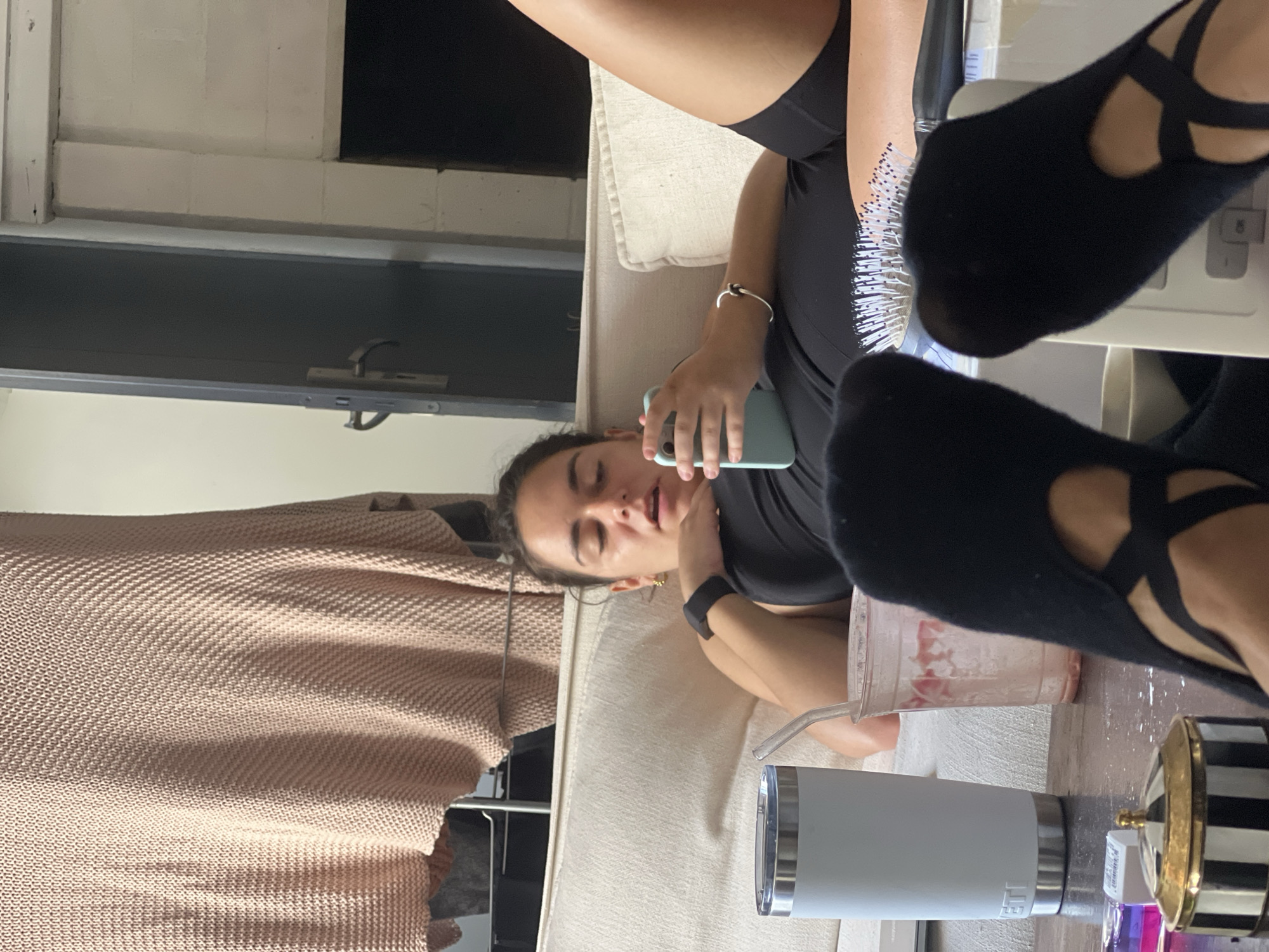 The image depicts an indoor scene, possibly a living room. A woman is lying on a light-colored sofa, holding a light blue smartphone. She is wearing a black athletic top and black shorts, with black socks on her feet. Her eyes are open and she appears to be looking at the phone. On the coffee table in front of the sofa, there is a light blue insulated tumbler, a clear plastic cup with a pink beverage and straw, and a black and white striped lidded container. In the background, a knitted throw is draped over what appears to be a shelf or rack. The overall lighting suggests it is daytime, but the ambient light level is moderate. There are no discernible features that place the location specifically in Paris, France.